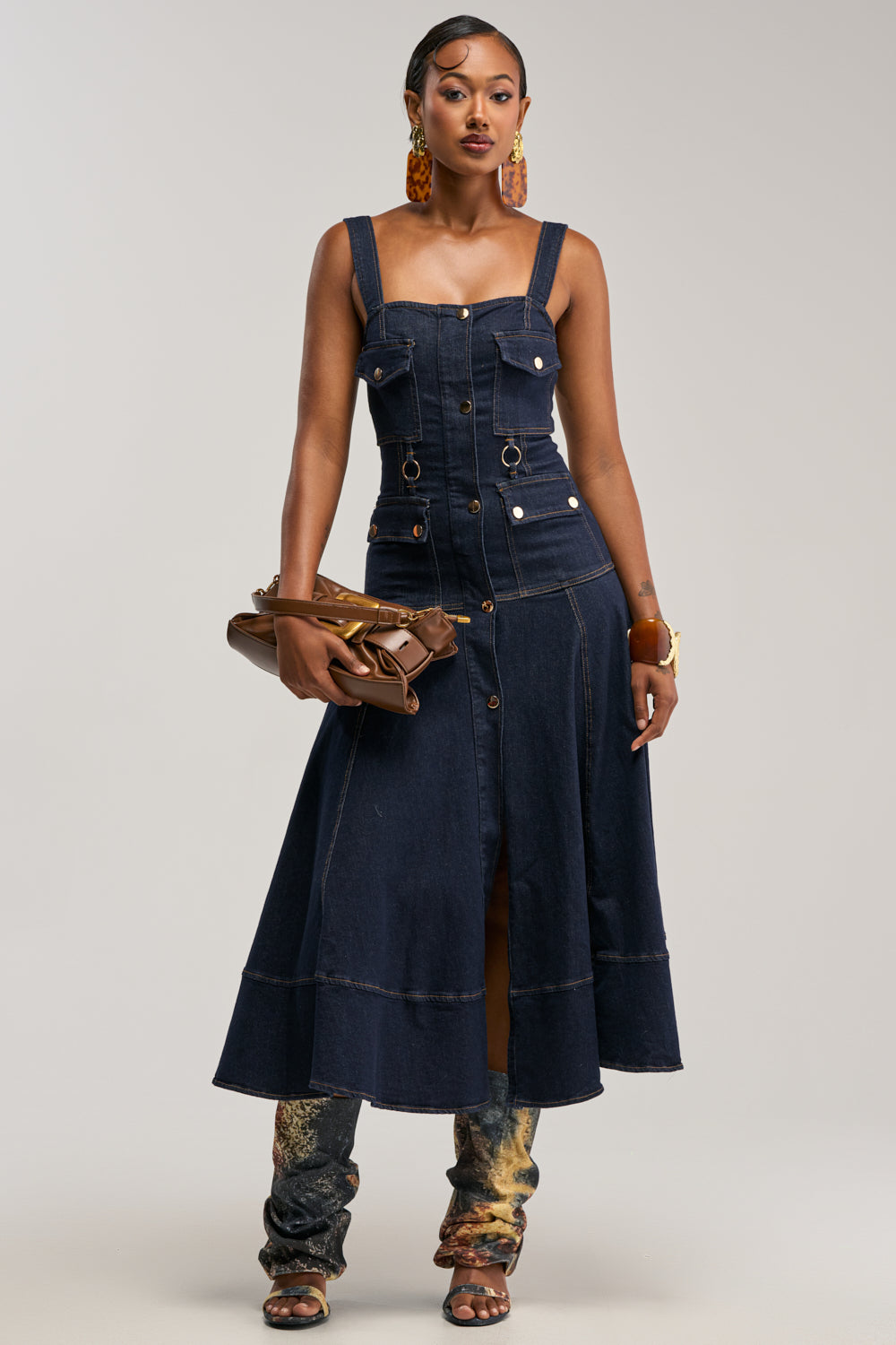 A woman exudes effortless chic and modern style in the WILDFLOWER DRIFT MIDI DRESS IN DARK BLUE DENIM—a sleeveless, utility-inspired design with front buttons. She completes her fashionable look with statement earrings, layered bracelets, a brown clutch, and patterned boots, striking a confident pose against a neutral background.