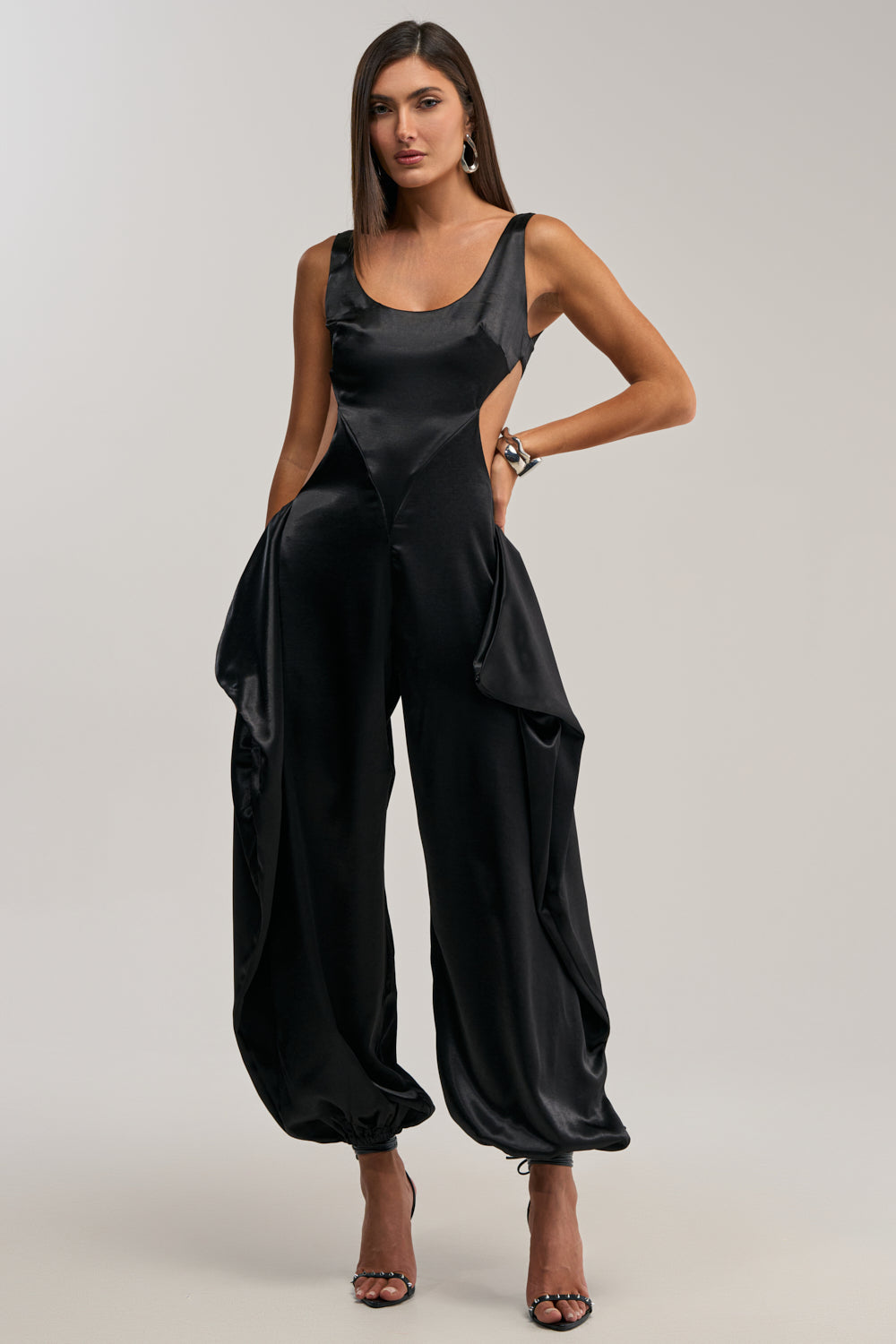 A woman models the A MOMENT IN SATIN JUMPSUIT: a sleeveless black satin jumpsuit with cut-out sides, draped wide legs gathered at the ankles, styled with open-toe heels, straight hair, hoop earrings, and a confident hand-on-hip pose.