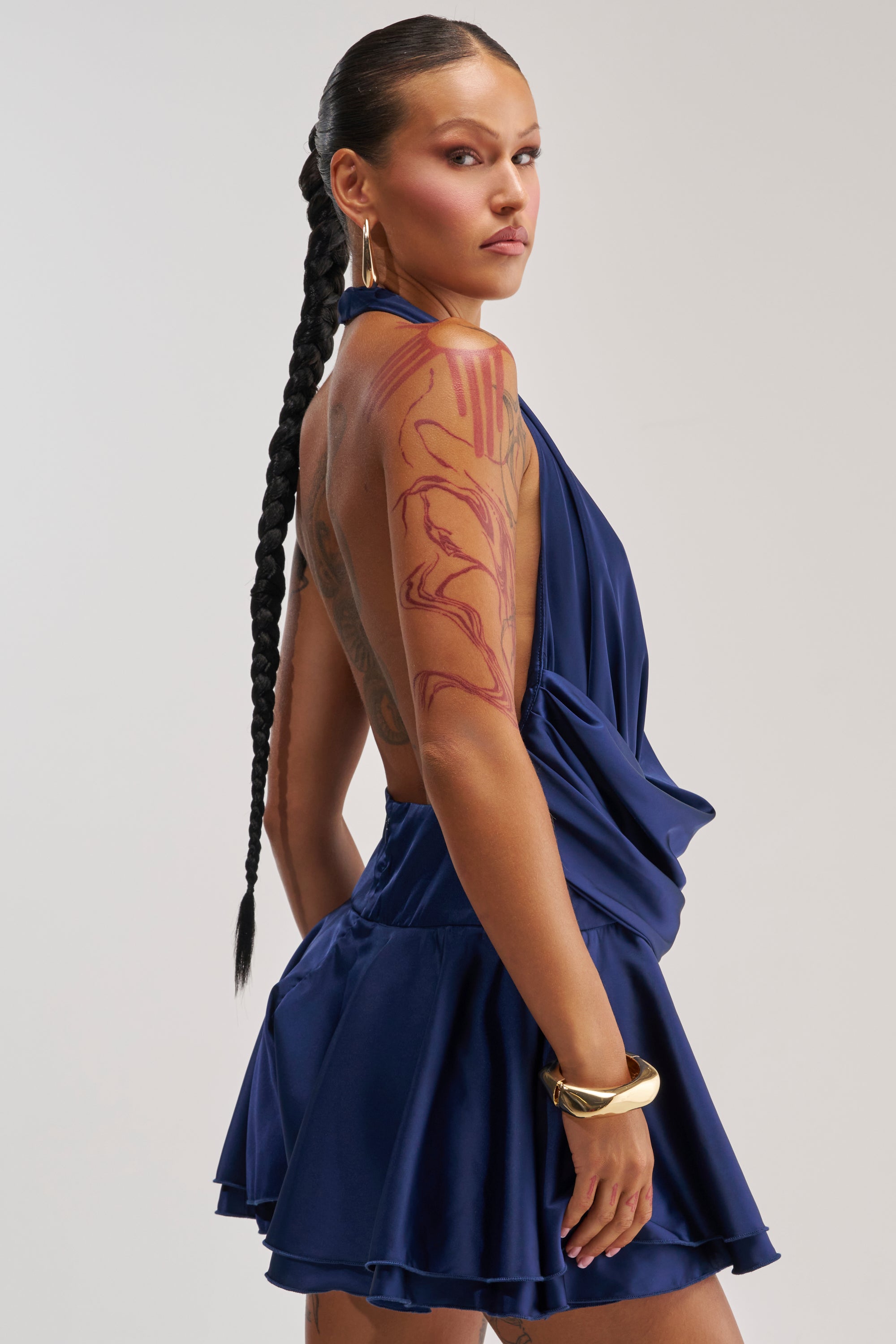 A person with long braided hair, gold hoop earrings, and a chunky gold bracelet poses confidently in the UNDERSTOOD THE ASSIGNMENT SATIN MINI DRESS IN NAVY—a blue sleeveless open-back halter mini dress. Their large abstract arm tattoo adds an edgy flair to this elegant look, making it a perfect choice for a stunning homecoming dress.
