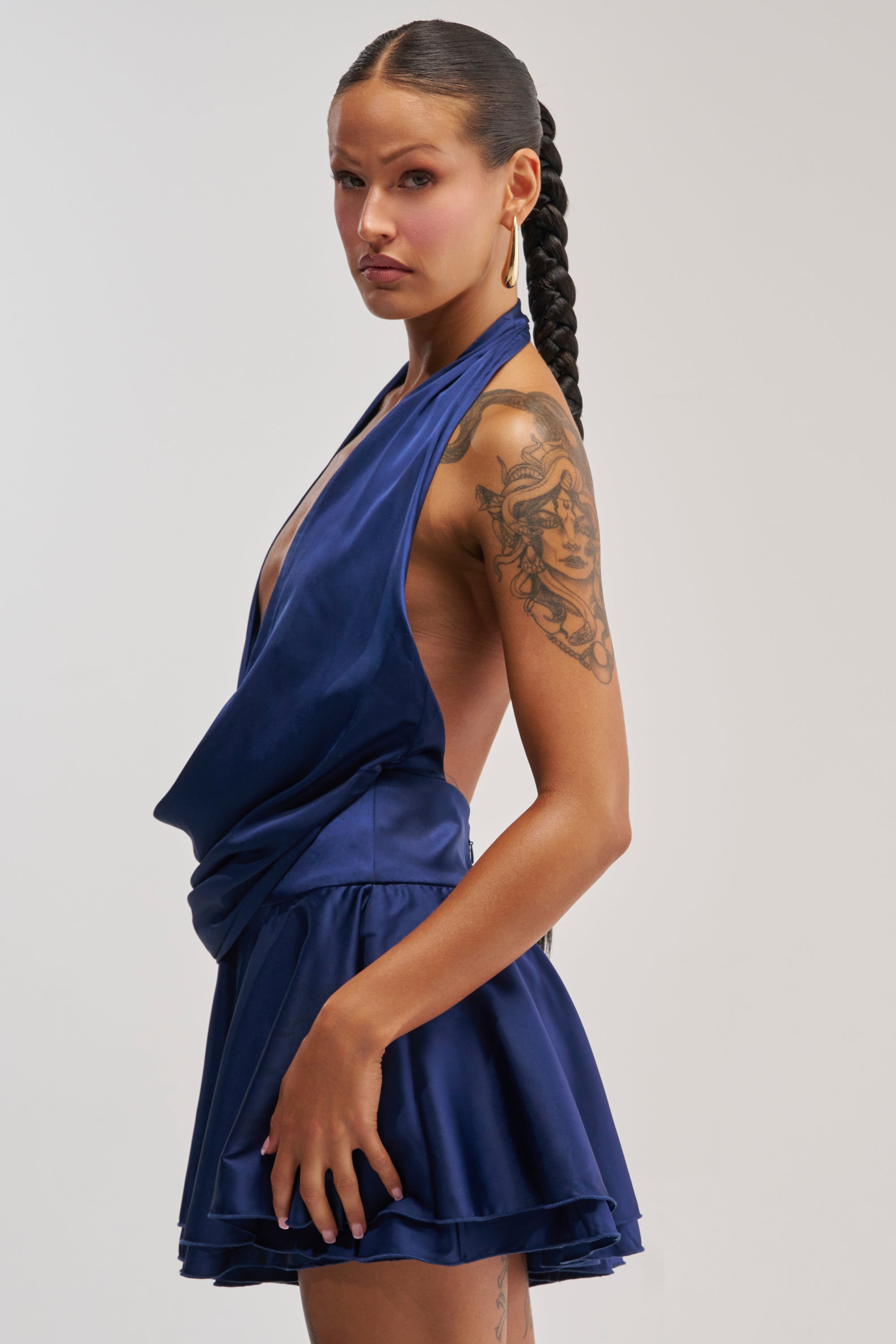 A woman with a long braid showcases the UNDERSTOOD THE ASSIGNMENT SATIN MINI DRESS IN NAVY—a sleeveless, backless homecoming look—paired with gold earrings that accentuate her tattooed upper arm, standing against a plain light background.