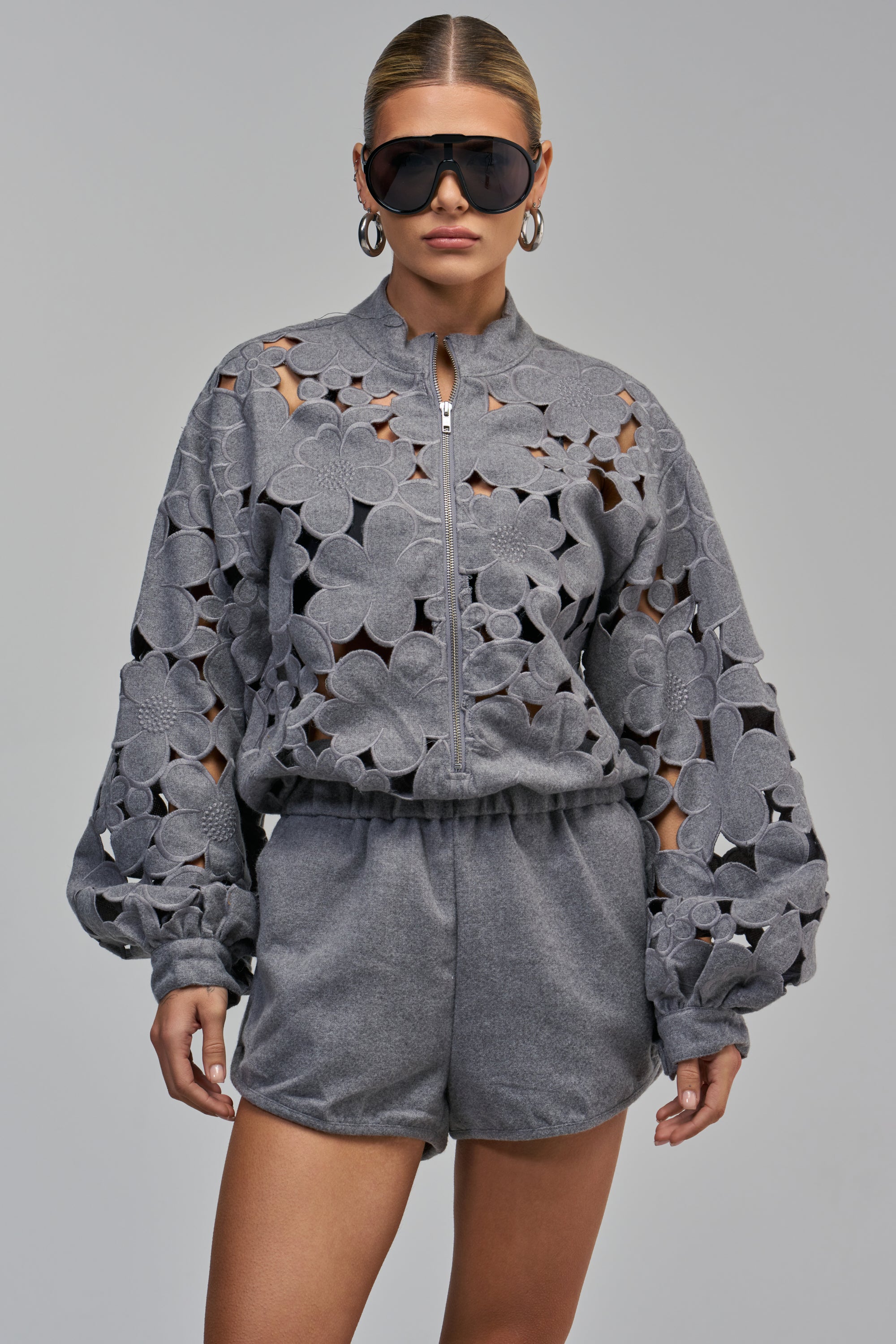 A person wearing the ATTENTION SEEKER FLORAL ROMPER, a grey floral cutout style with a zip-up front, long loose sleeves, and large floral patterns throughout the bodice and sleeves.