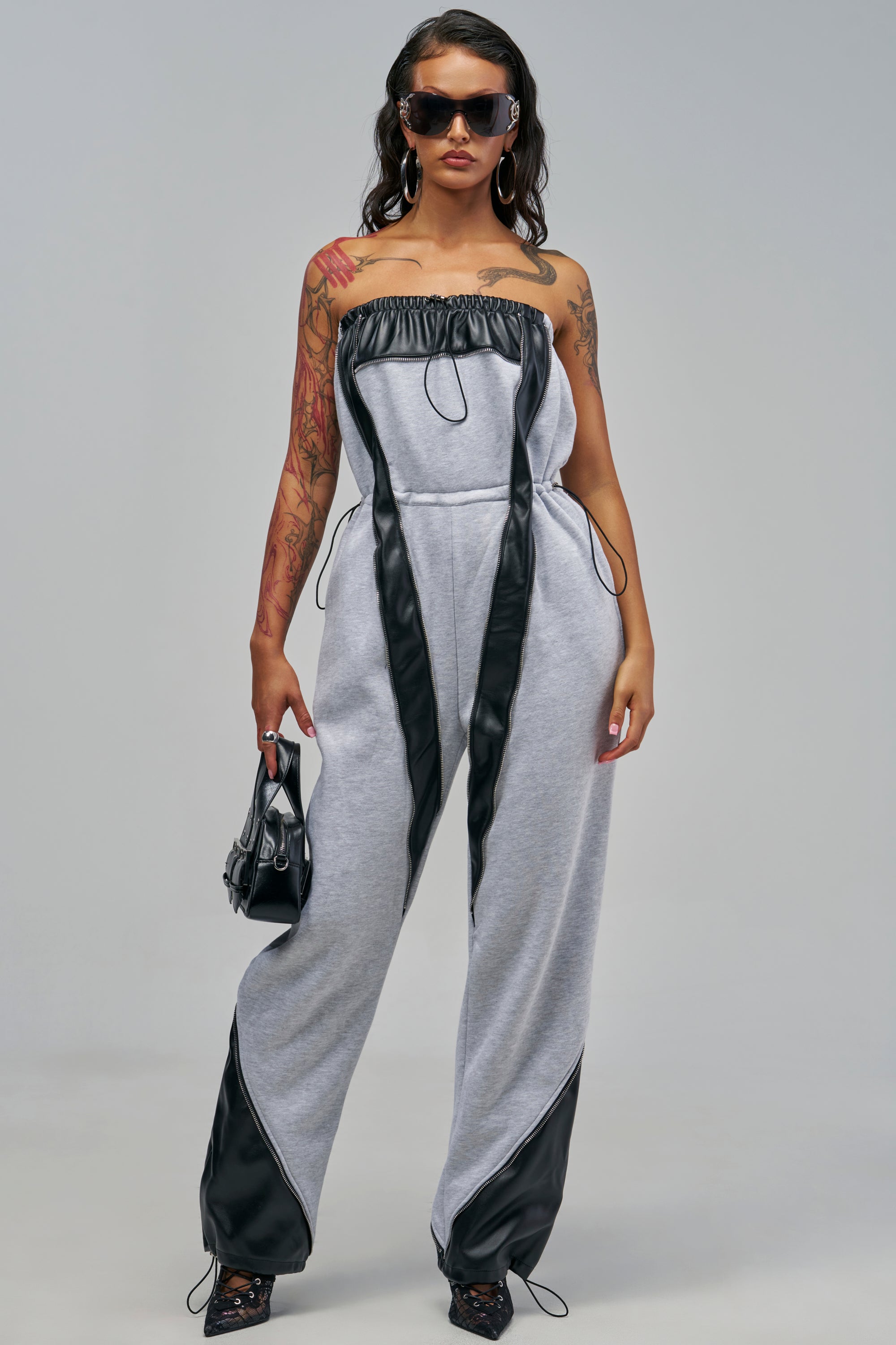 A person wearing the grey faux leather SHE RUNS IT STRAPLESS JUMPSUIT with black trim accents, black sunglasses, and holding a small black handbag.