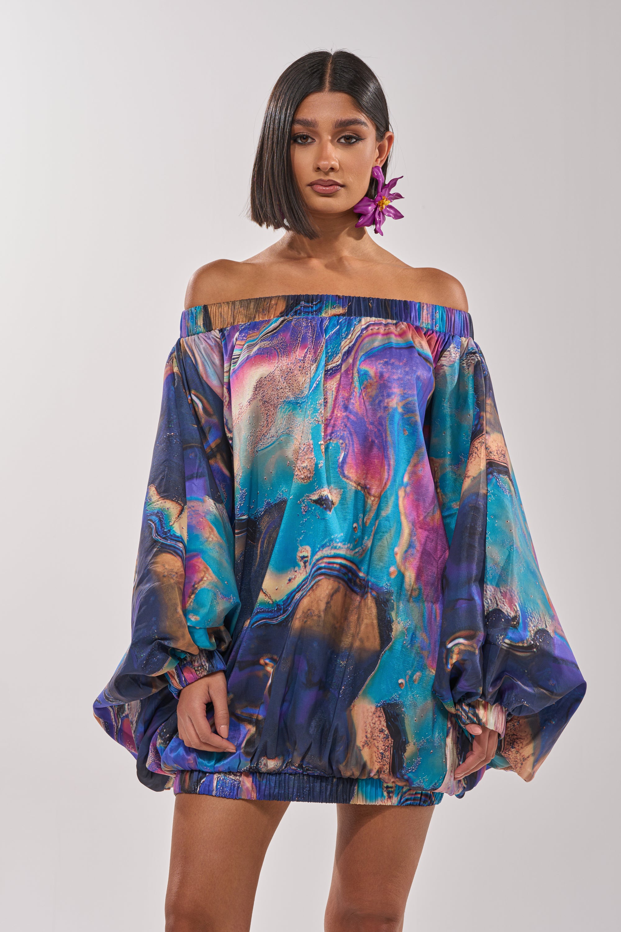 A woman with a sleek bob wears the OUT FOR BRUNCH PATTERNED MINI DRESS, featuring abstract swirls, an off-the-shoulder neckline, and long balloon sleeves. She accessorizes with large purple flower earrings against a plain background. 0