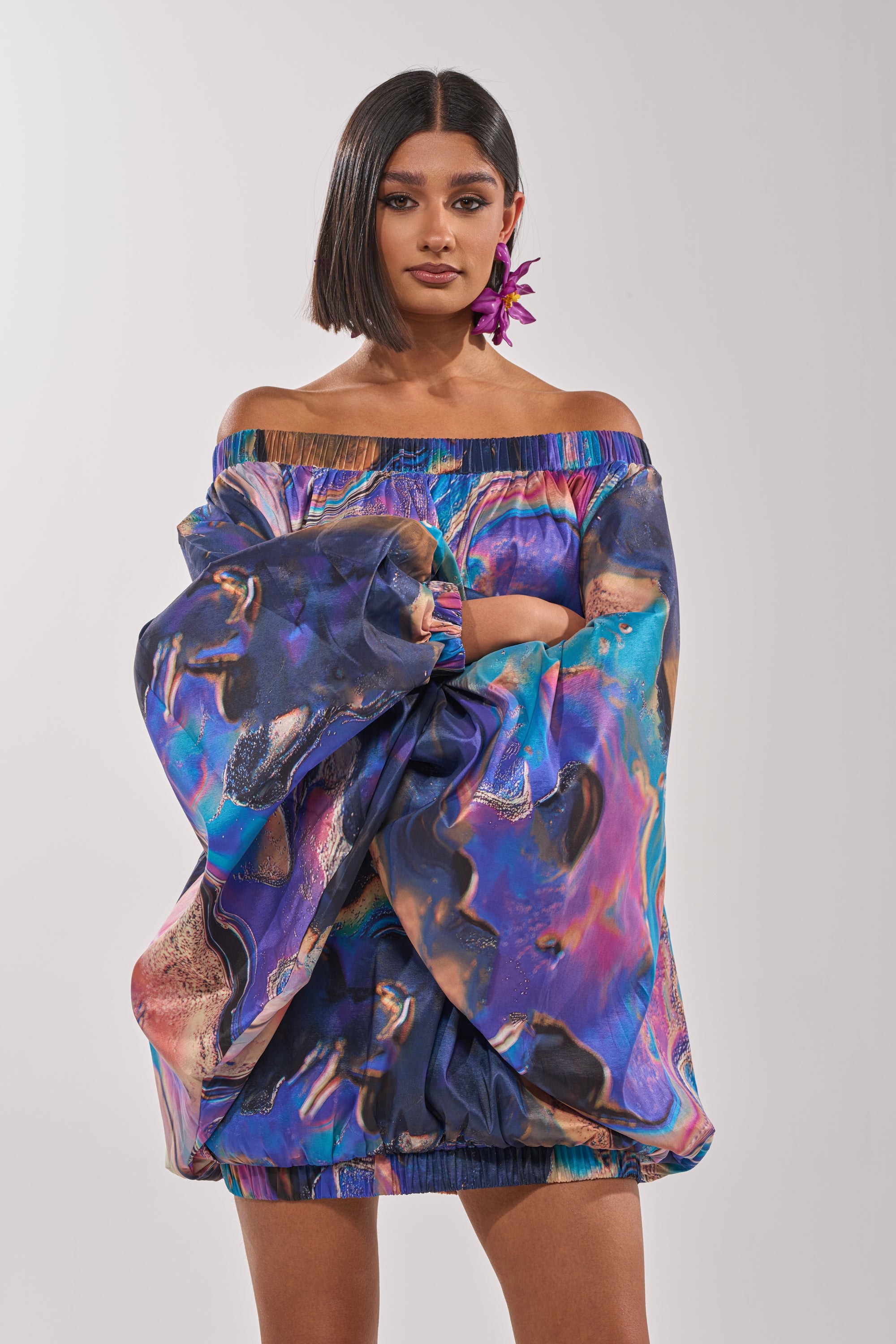 A woman with a sleek bob models the OUT FOR BRUNCH PATTERNED MINI DRESS, showcasing its colorful abstract swirl print and voluminous balloon sleeves, styled with a large purple flower earring against a plain light background. 2