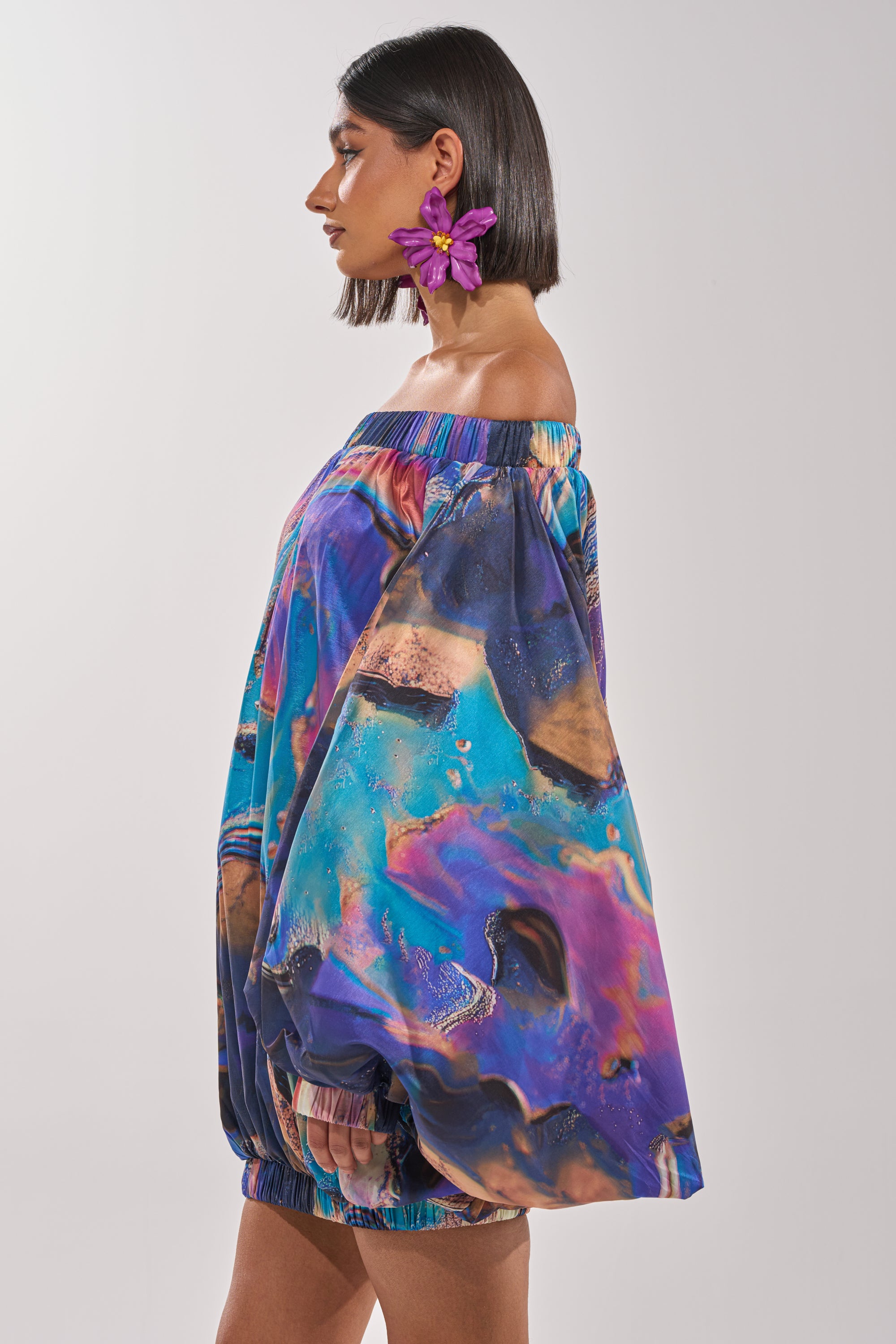 A woman with straight dark hair and a purple flower earring models the OUT FOR BRUNCH PATTERNED MINI DRESS, an off-the-shoulder mini dress with a bold blue, purple, and orange abstract pattern. She poses in profile against a plain background. 3
