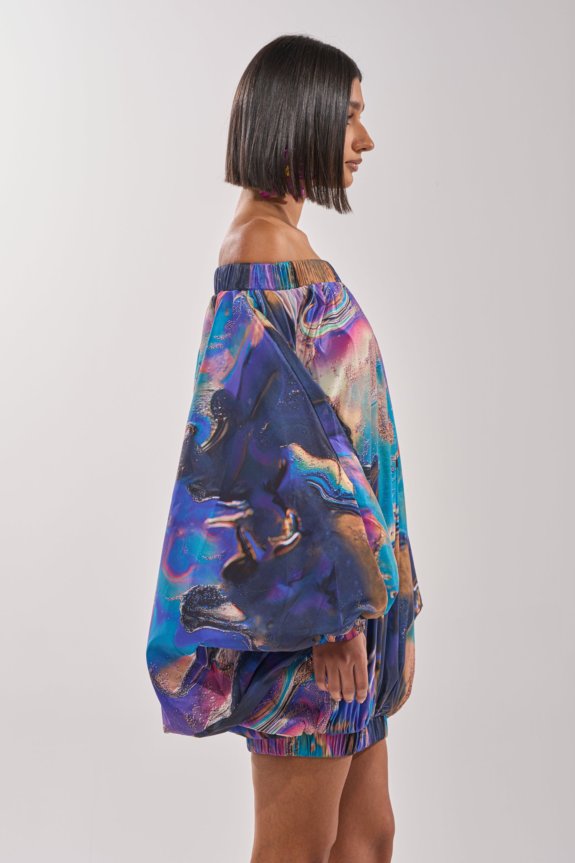 A woman with a straight bob stands in profile, wearing the OUT FOR BRUNCH PATTERNED MINI DRESS—an off-the-shoulder, balloon sleeve mini dress with vibrant blue and purple abstract swirls. The background is plain and light-colored. 4