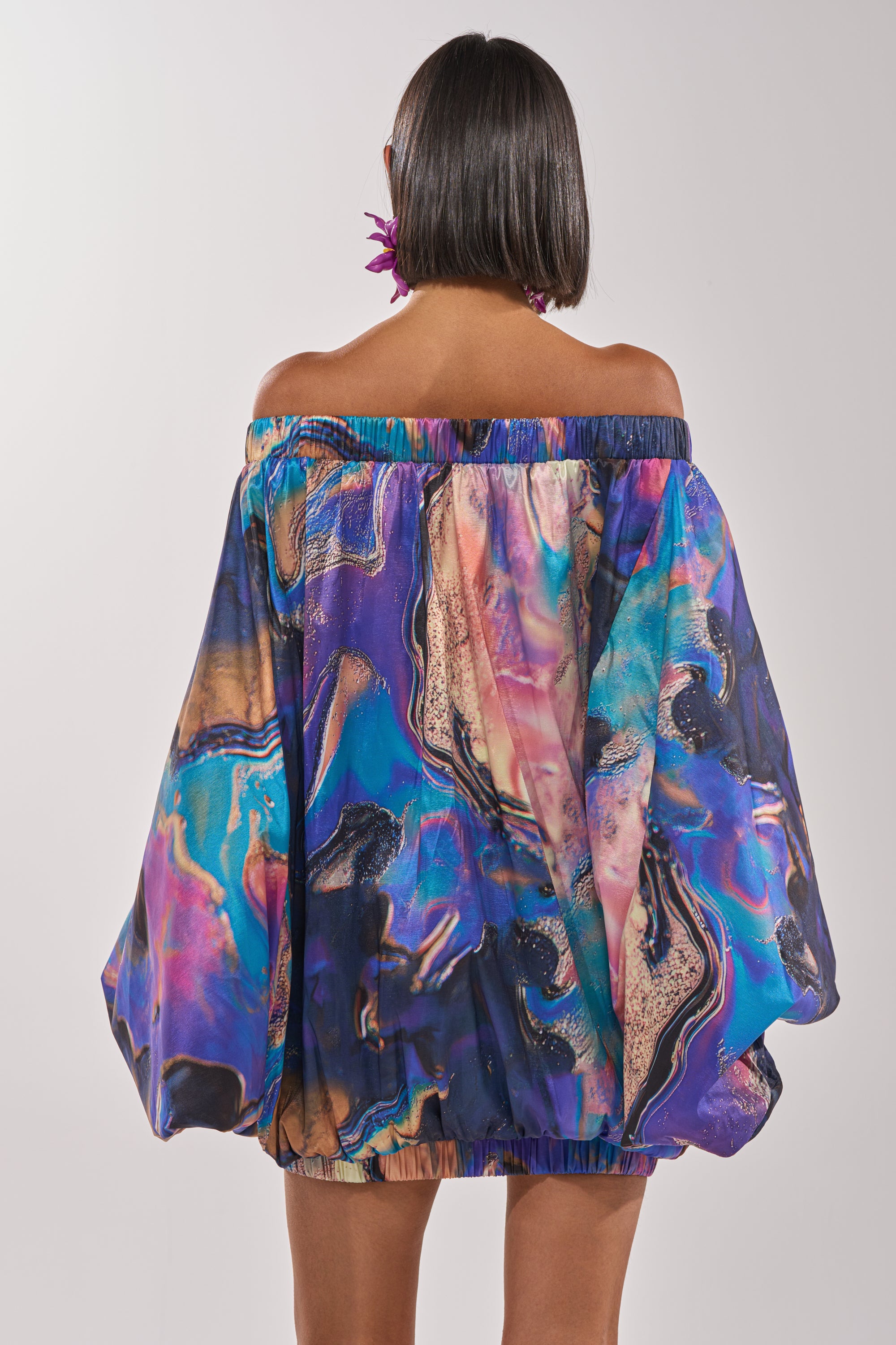 A person with a bob haircut, seen from behind, wears the OUT FOR BRUNCH PATTERNED MINI DRESS—an off-the-shoulder balloon sleeve dress with a vibrant abstract swirl pattern in blue, purple, pink, and gold tones. 5