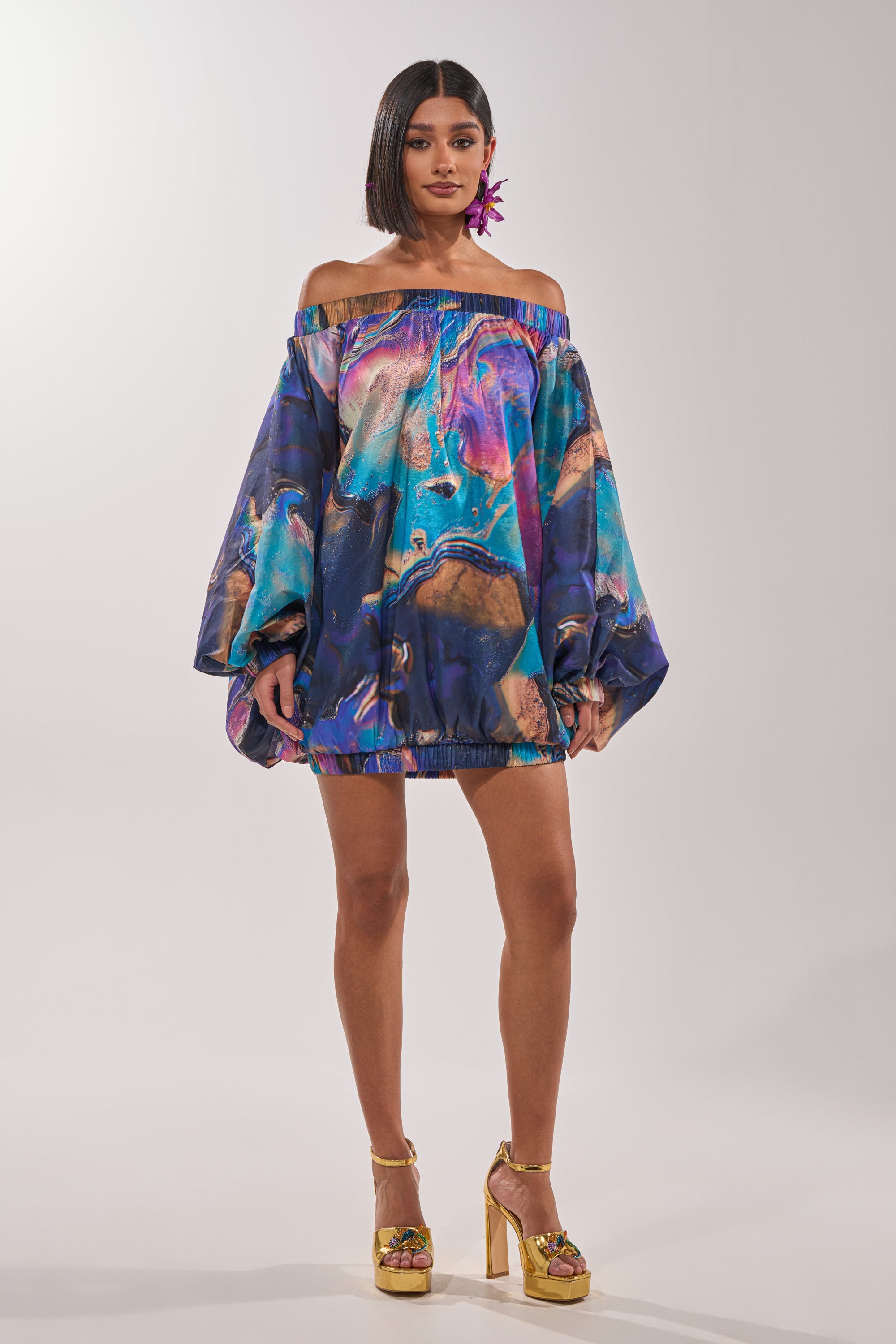 A woman wearing the OUT FOR BRUNCH PATTERNED MINI DRESS, featuring off-the-shoulder balloon sleeves and a vibrant abstract swirl print, stands facing forward. She pairs it with a bob haircut, statement earrings, and gold high-heeled sandals. 6