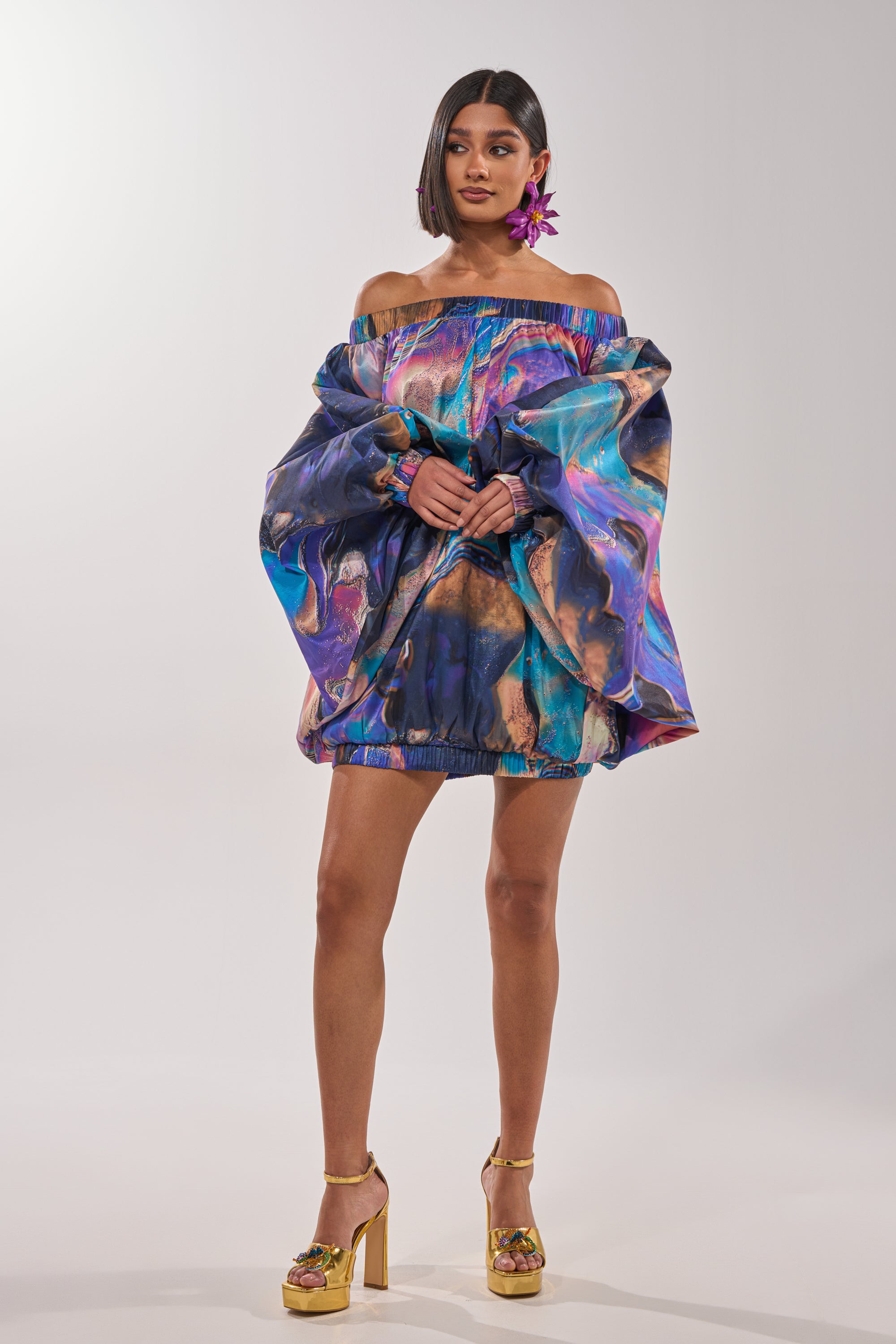 A woman poses against a plain background wearing the OUT FOR BRUNCH PATTERNED MINI DRESS—an off-the-shoulder style with balloon sleeves and a vibrant blue and purple abstract swirl print—paired with gold heels and a bold purple flower earring. 7