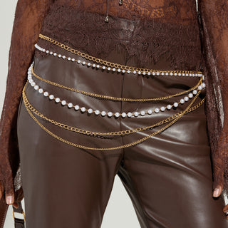 A person wearing a sheer, long-sleeved brown lace top and brown leather pants adorned with the PEARL PLEASE LAYERED CHAIN BELT, featuring adjustable lobster clasp closures.