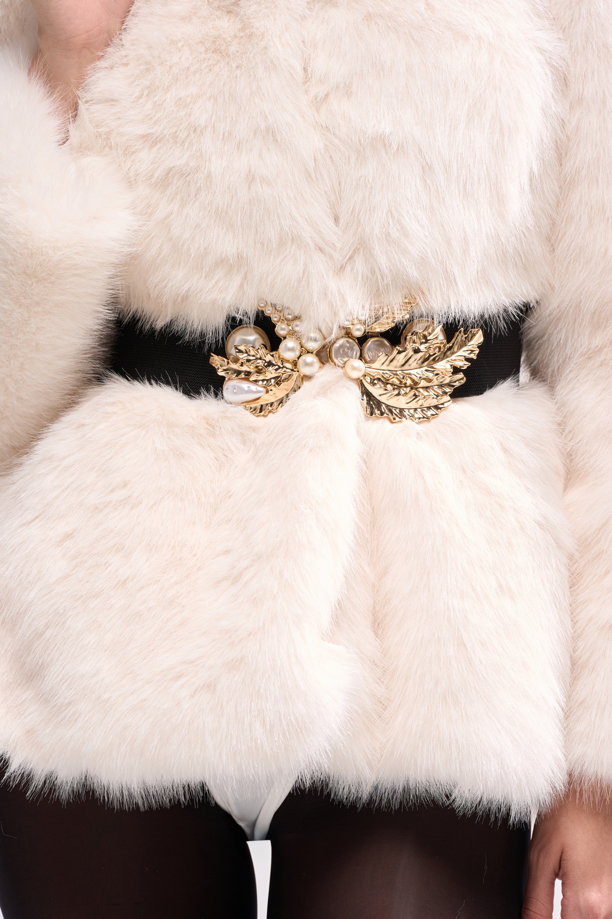 A person wears a white faux fur coat with the EXQUISITE TASTE STRETCH BELT, which features a gold leaf buckle and pearl embellishments. Only the torso and part of one arm are visible.
