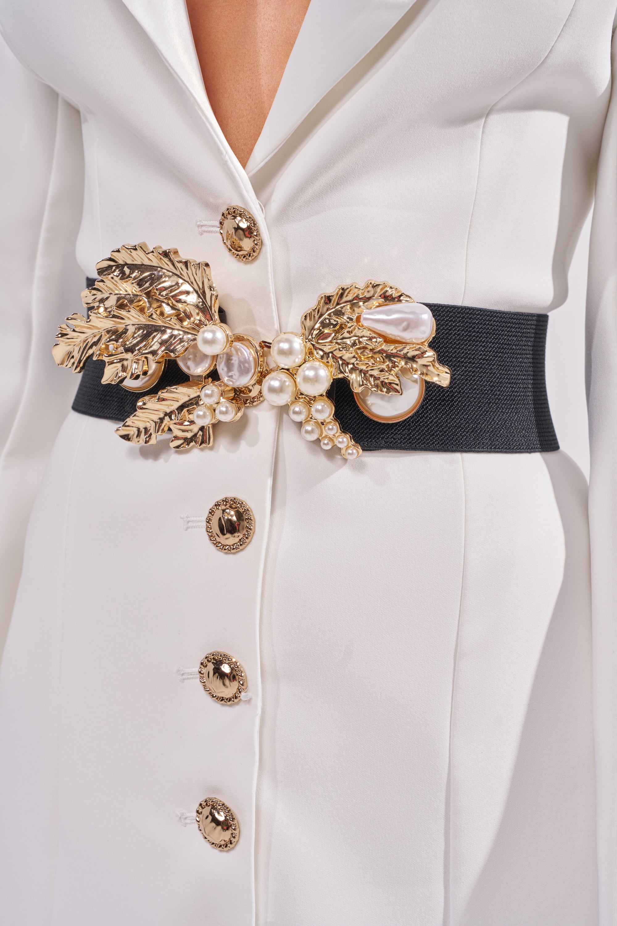 A close-up of a person wearing a white blazer with gold buttons, styled with the EXQUISITE TASTE STRETCH BELT—a black waist belt featuring a gold leaf buckle, pearls, and decorative accents at the waist. 1