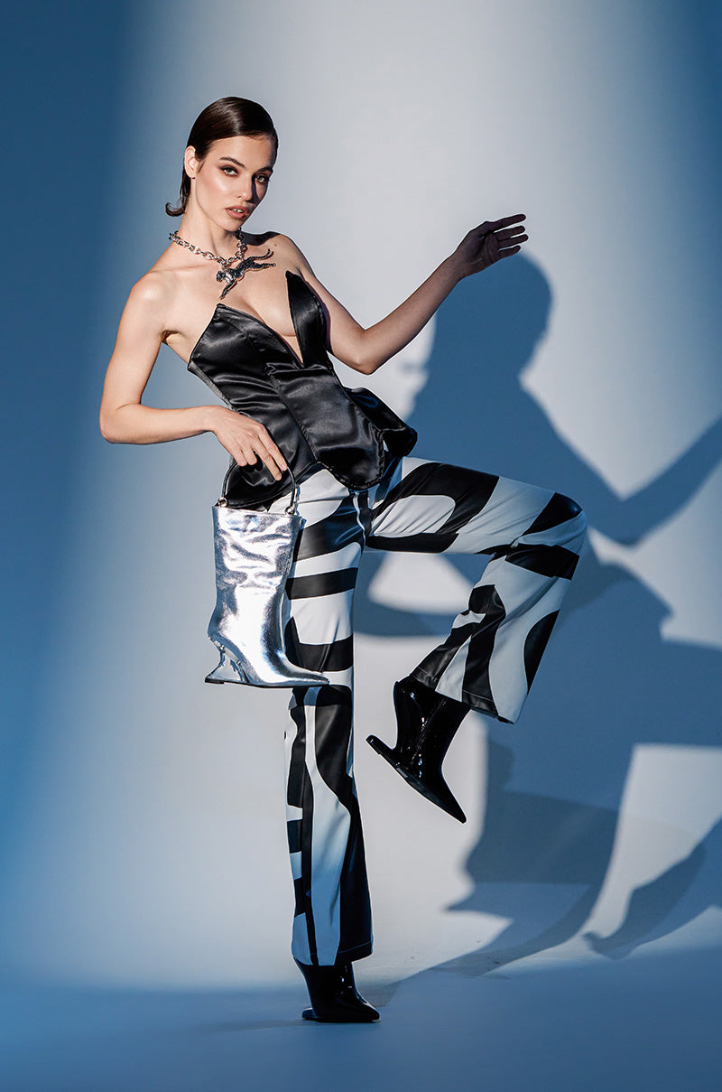 A woman poses confidently in GROOVE ON FAUX LEATHER FLARE TROUSER with a strapless top, shiny boots, metallic handbag, and chunky necklace against dramatic lighting and shadows.