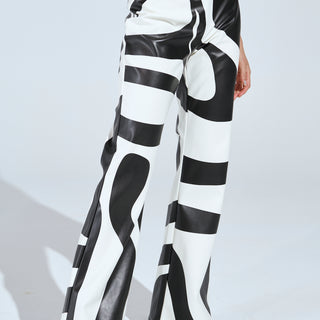 A person wears the GROOVE ON FAUX LEATHER FLARE TROUSER, featuring a black and white swirl pattern. They pair it with black pointed shoes adorned with silver studs. The flared leg silhouette enhances the dramatic, artistic design, while lighting casts a shadow on the white background.