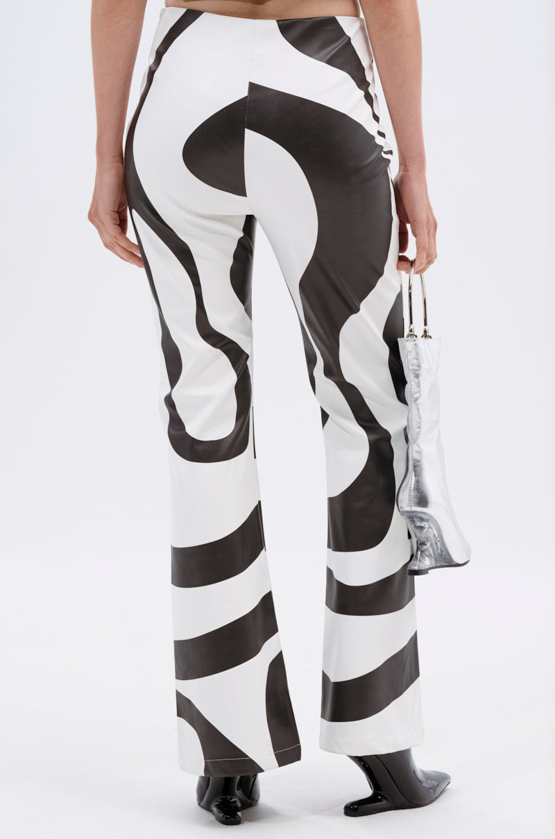 A person stands facing away on a plain white background, wearing GROOVE ON FAUX LEATHER FLARE TROUSER with a black-and-white swirl print, black pointed shoes, and holding a small metallic silver boot-shaped purse.