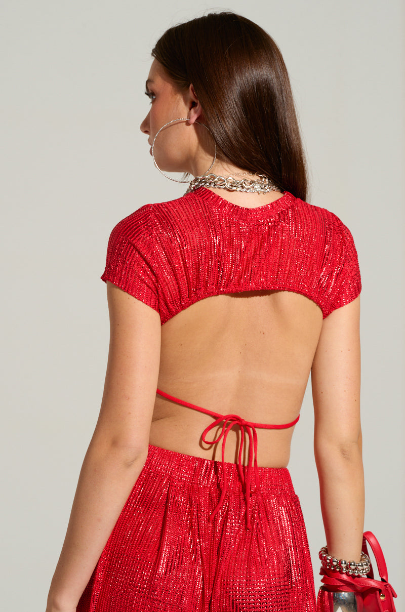A woman is seen from the back, wearing a sparkly red "Just Like Magic Metallic Open Back Top" from AKIRA Label, tied with a string. She accessorizes with hoop earrings, a silver necklace, bracelets, and is holding a red handbag. Her straight brown hair cascades past her shoulders in a chic cropped silhouette.