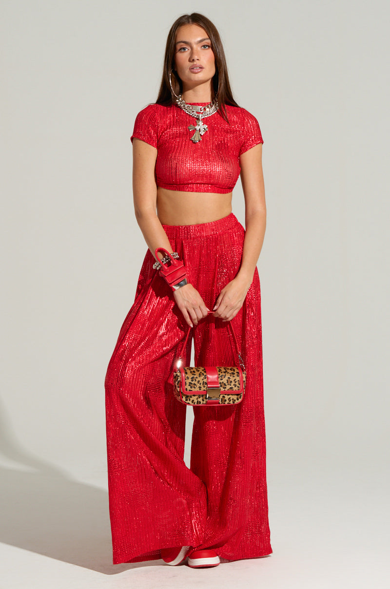 A person poses against a neutral background, showcasing the JUST LIKE MAGIC METALLIC OPEN BACK TOP IN RED along with matching shimmering wide-leg pants. They carry a small leopard-print handbag, adorned with multiple bracelets, their long hair flowing with a confident expression.