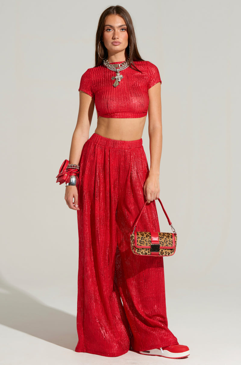 A woman stands wearing the JUST LIKE MAGIC METALLIC OPEN BACK TOP IN RED as part of a bright, textured two-piece outfit with a cropped silhouette. She accessorizes with a statement necklace, stack of bracelets, and a small handbag, complemented by her red shoes. The background is plain.