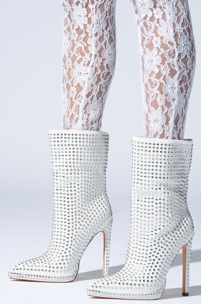 A pair of legs wearing white lace tights with AZALEA WANG Jubal White Embellished Bootie, featuring sparkly faux leather and stiletto heels, adorned with rhinestones. The boots showcase pointed toes against a plain background.