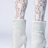 A pair of legs wearing white lace tights with AZALEA WANG Jubal White Embellished Bootie, featuring sparkly faux leather and stiletto heels, adorned with rhinestones. The boots showcase pointed toes against a plain background.