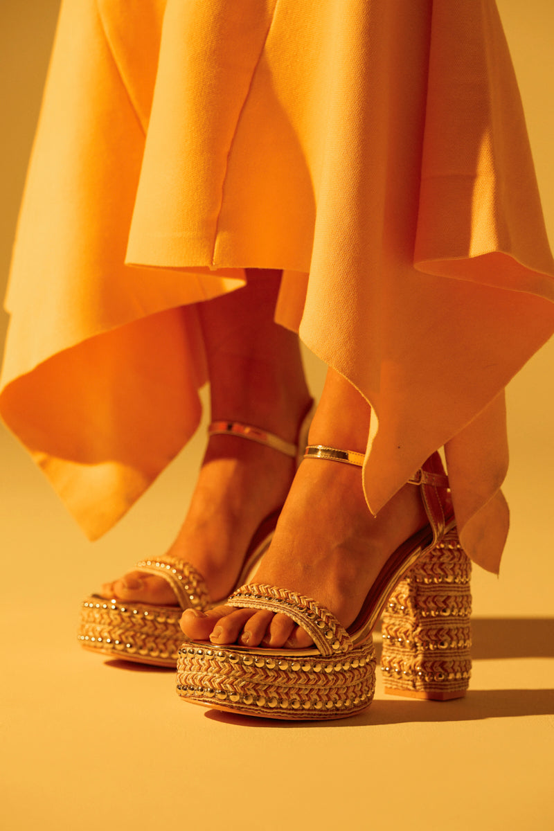 A person wearing AZALEA WANG JUDSON GOLD RAFFIA AND GEMS PLATFORM SANDAL, which features intricate woven designs, a chunky heel, thick platforms, and elegant gold metallic studs. They stand in a flowing asymmetrical orange dress on a warm golden-hued surface.