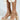 A close-up shows someone wearing AZALEA WANG JUDSON GOLD RAFFIA AND GEMS PLATFORM SANDALS, featuring intricate braids, chunky heels, gold metallic studs, and thick patterned platform soles. Their outfit has circular textured decorations on a plain light background.