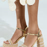 A close-up shows someone wearing AZALEA WANG JUDSON GOLD RAFFIA AND GEMS PLATFORM SANDALS, featuring intricate braids, chunky heels, gold metallic studs, and thick patterned platform soles. Their outfit has circular textured decorations on a plain light background.