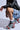 Squatting, a person wears a black leather jacket and denim shorts with cross patches while their AZALEA WANG Julien Blue Multi Chunky Bootie features a plaid textile upper embellished with rhinestones, highlighting the outfit.