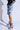 A person wearing a black leather jacket and denim shorts with black cross patterns showcases the AZALEA WANG JULIEN BLUE MULTI CHUNKY BOOTIE featuring a colorful plaid textile upper. The background is plain white.