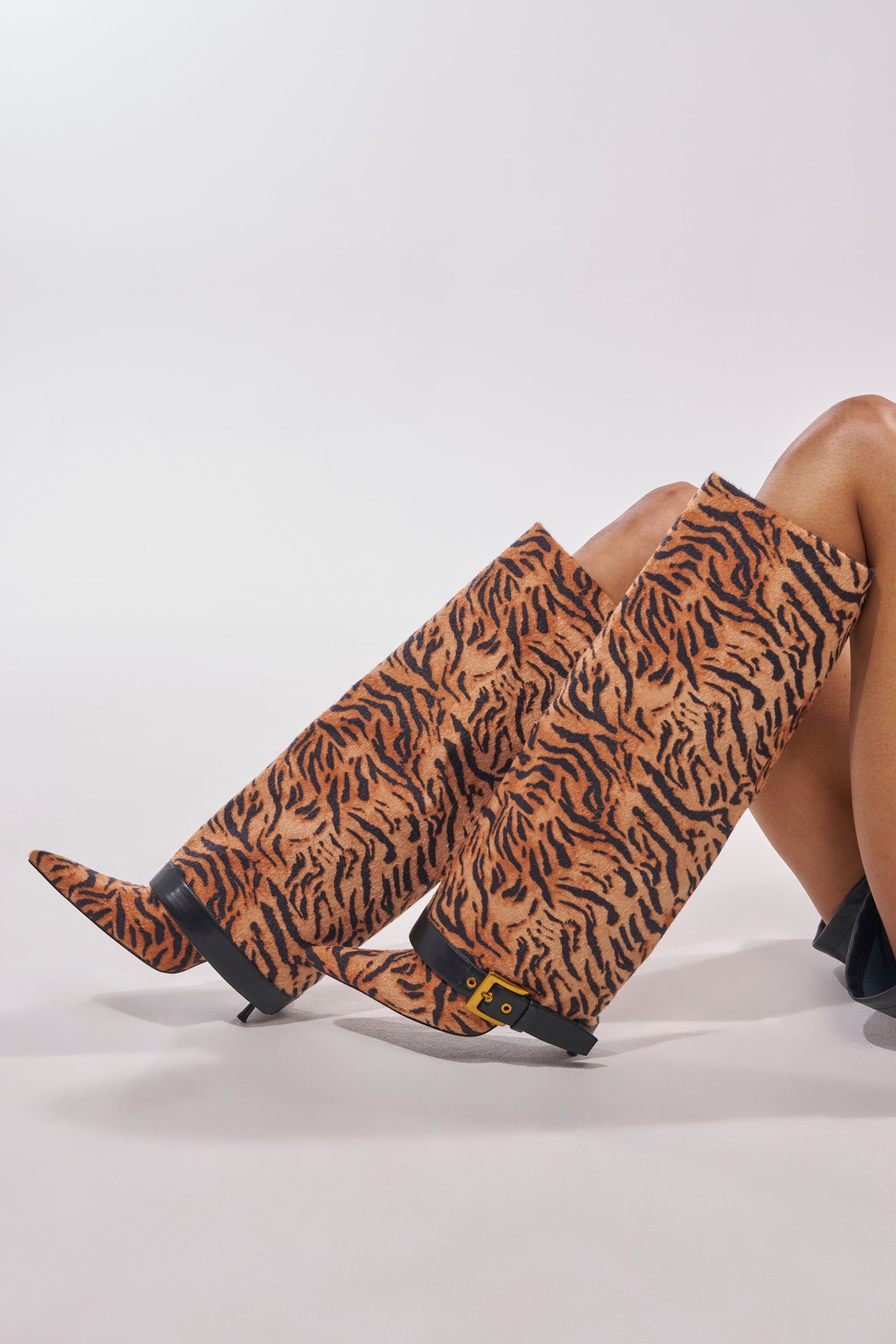 A person models the AZALEA WANG JUST TIGER STILETTO FOLD OVER BOOT—tiger print, knee-high with black trim and gold buckle—legs extended on a plain light background; bold statement boots for standout style. 0