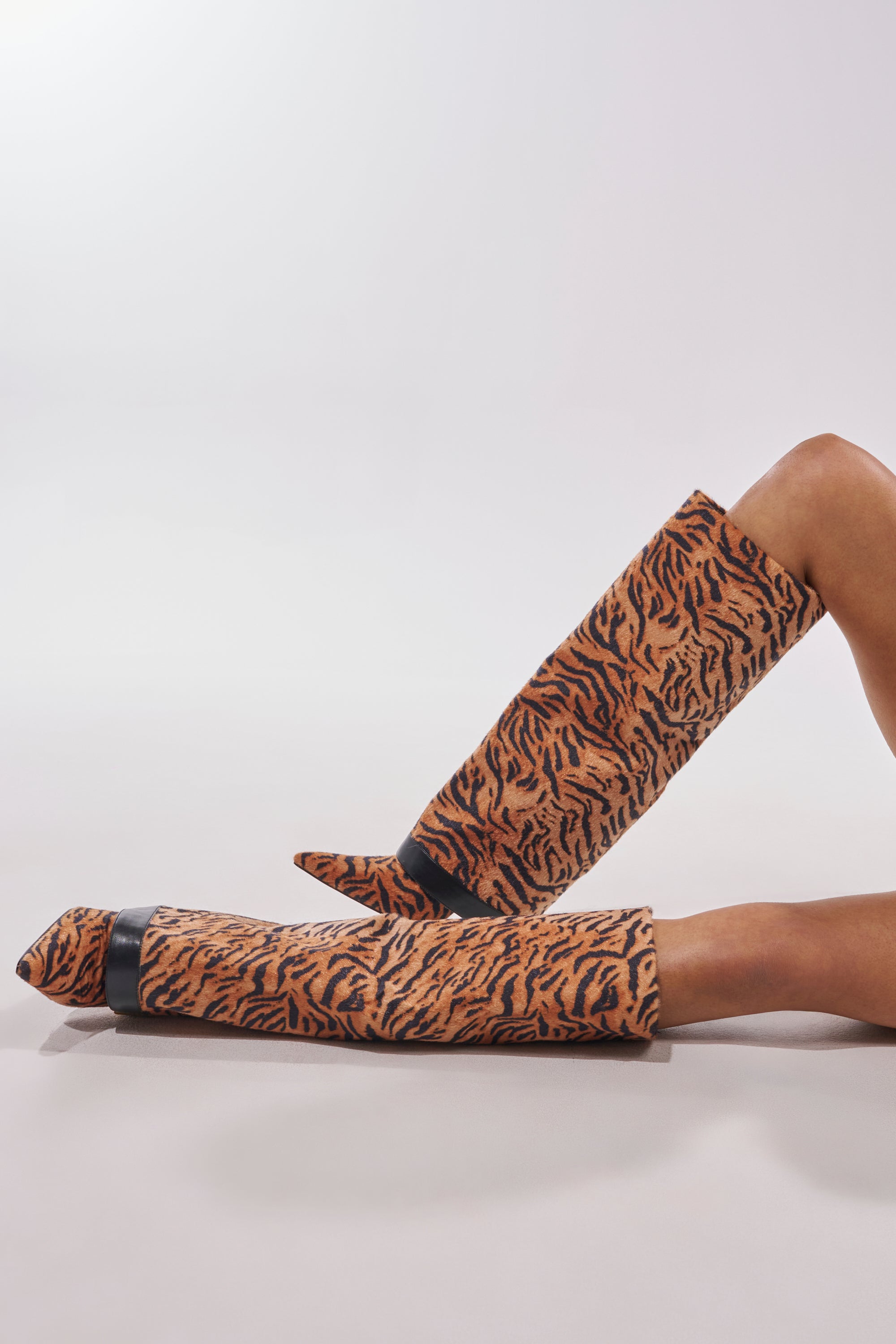 Two legs wearing the AZALEA WANG JUST TIGER STILETO FOLD OVER BOOT—tiger print knee-high boots with stiletto heels—rest on a light, neutral background. 1