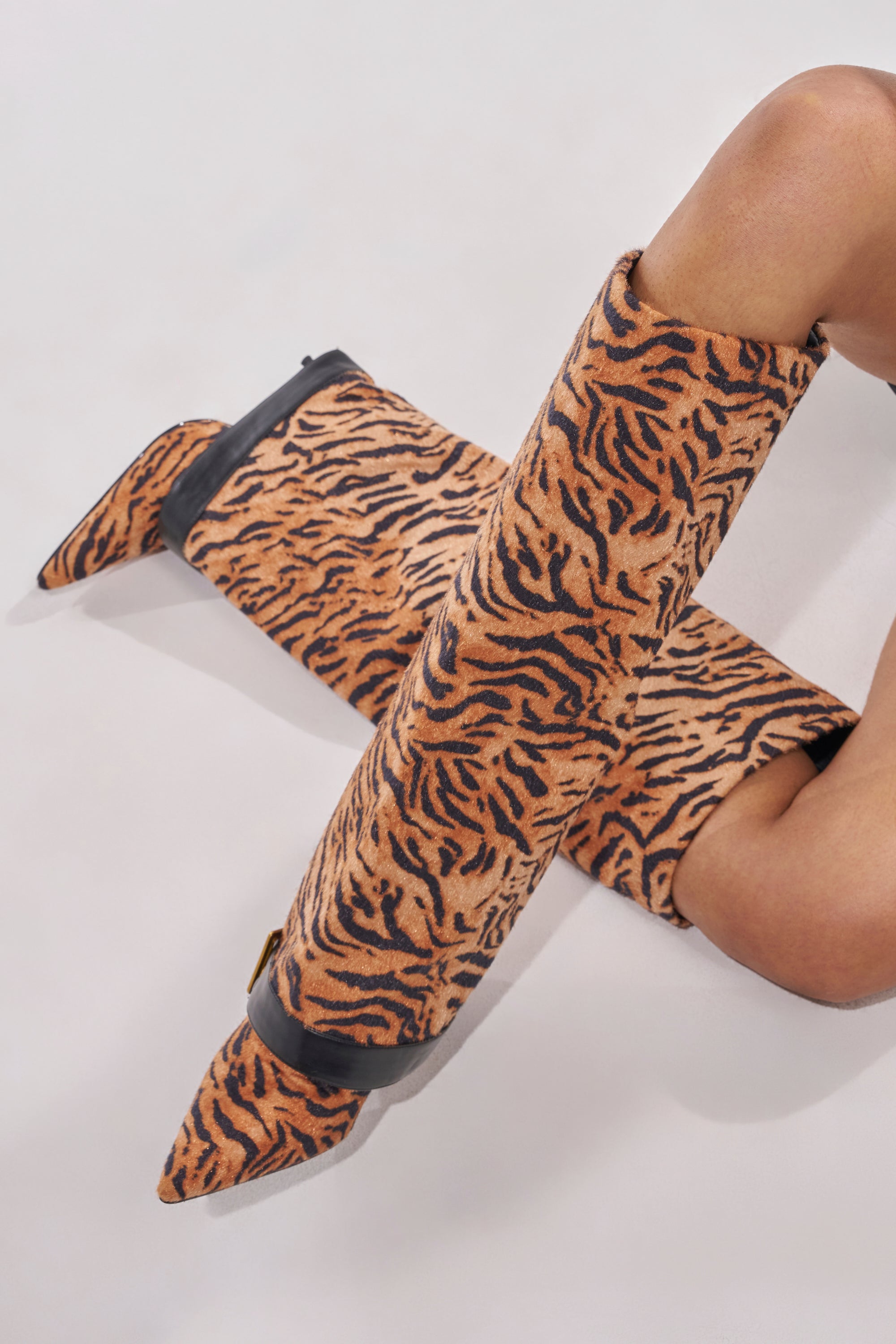 A person wearing AZALEA WANG JUST TIGER STILETTO FOLD OVER BOOTS with a bold tiger print and pointed toe sits on a white surface, one leg crossed over the other. 2