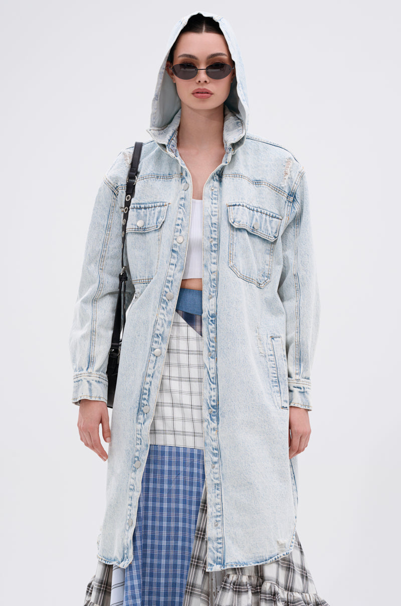 Wearing the ZOEY HOODED DENIM JACKET over a white top and plaid skirt, paired with dark sunglasses and a black shoulder bag, a person stands against a plain light background.