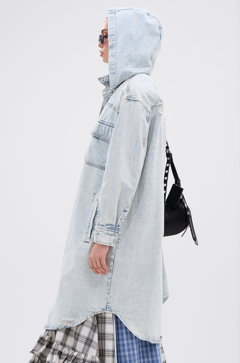 A person stands sideways in the ZOEY HOODED DENIM JACKET in light blue, paired with sunglasses, a black shoulder bag, and a layered plaid skirt against a plain white background.