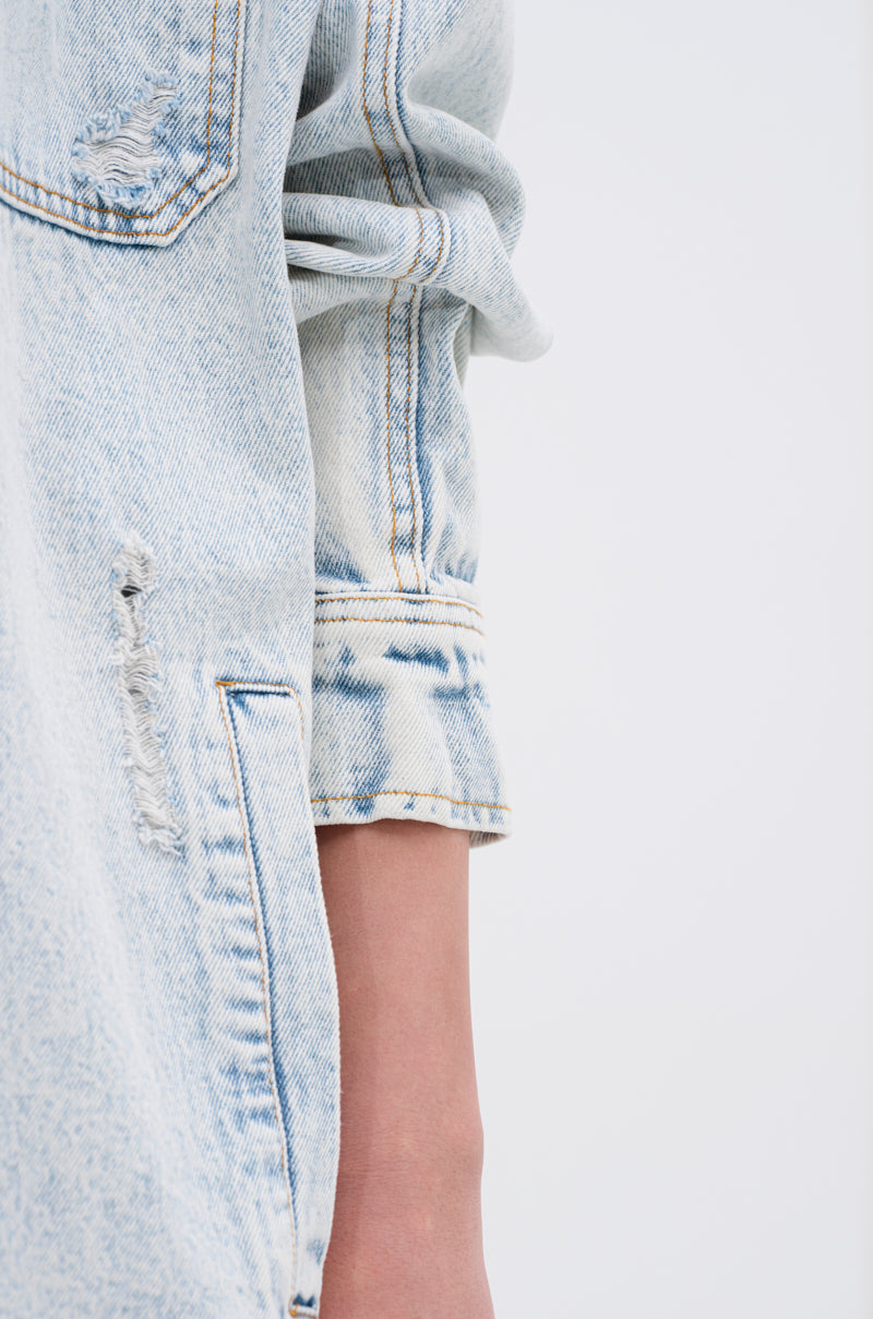 A close-up of a person wearing the ZOEY HOODED DENIM JACKET, featuring an oversized fit, light blue distressed denim, and rolled-up sleeves, with part of their arm visible against a plain light background.