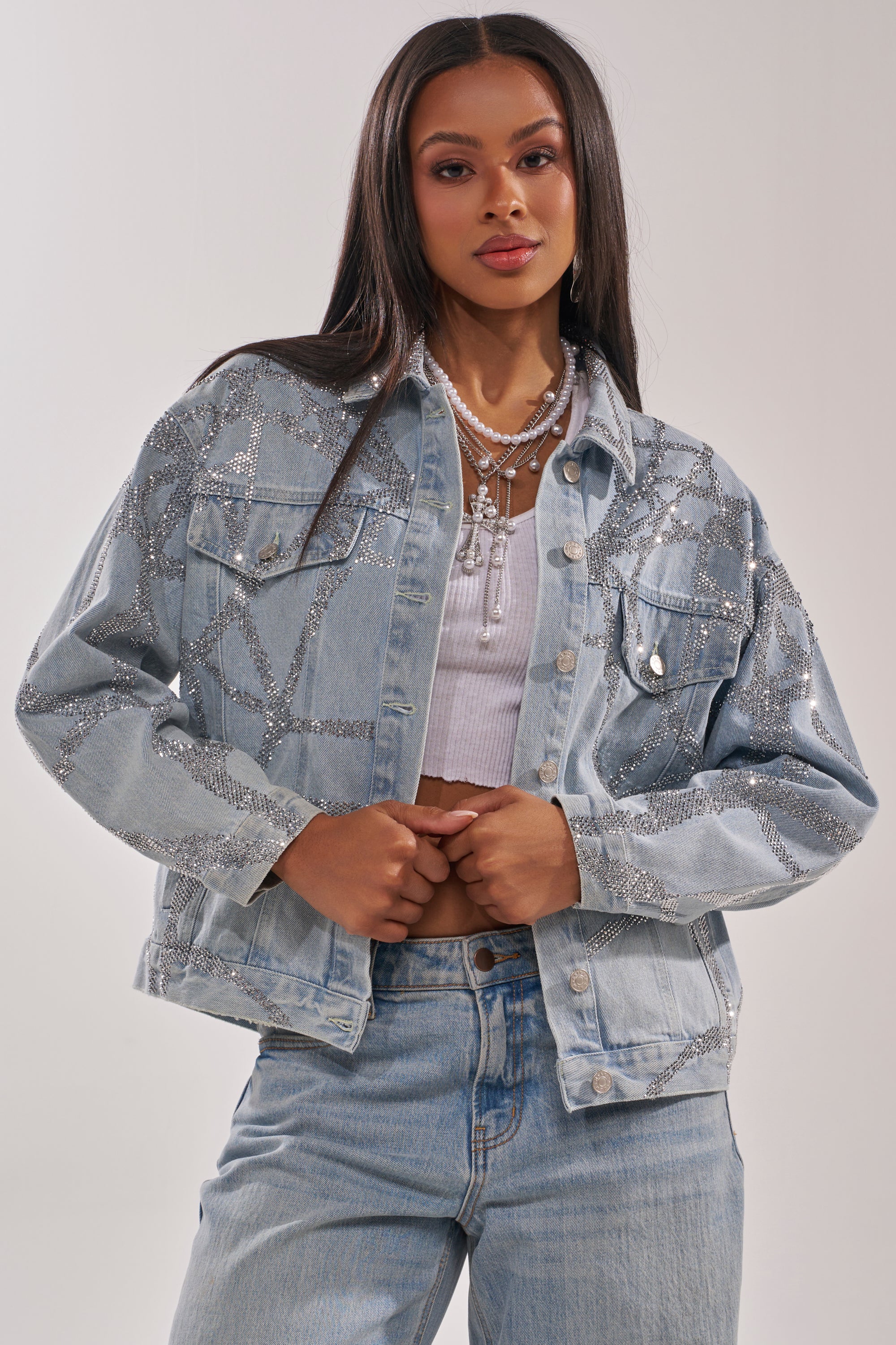 A woman stands against a plain backdrop wearing the TWILIGHT ZONE RHINESTONE DENIM JACKET over a white crop top and matching jeans, accessorized with layered necklaces. 0