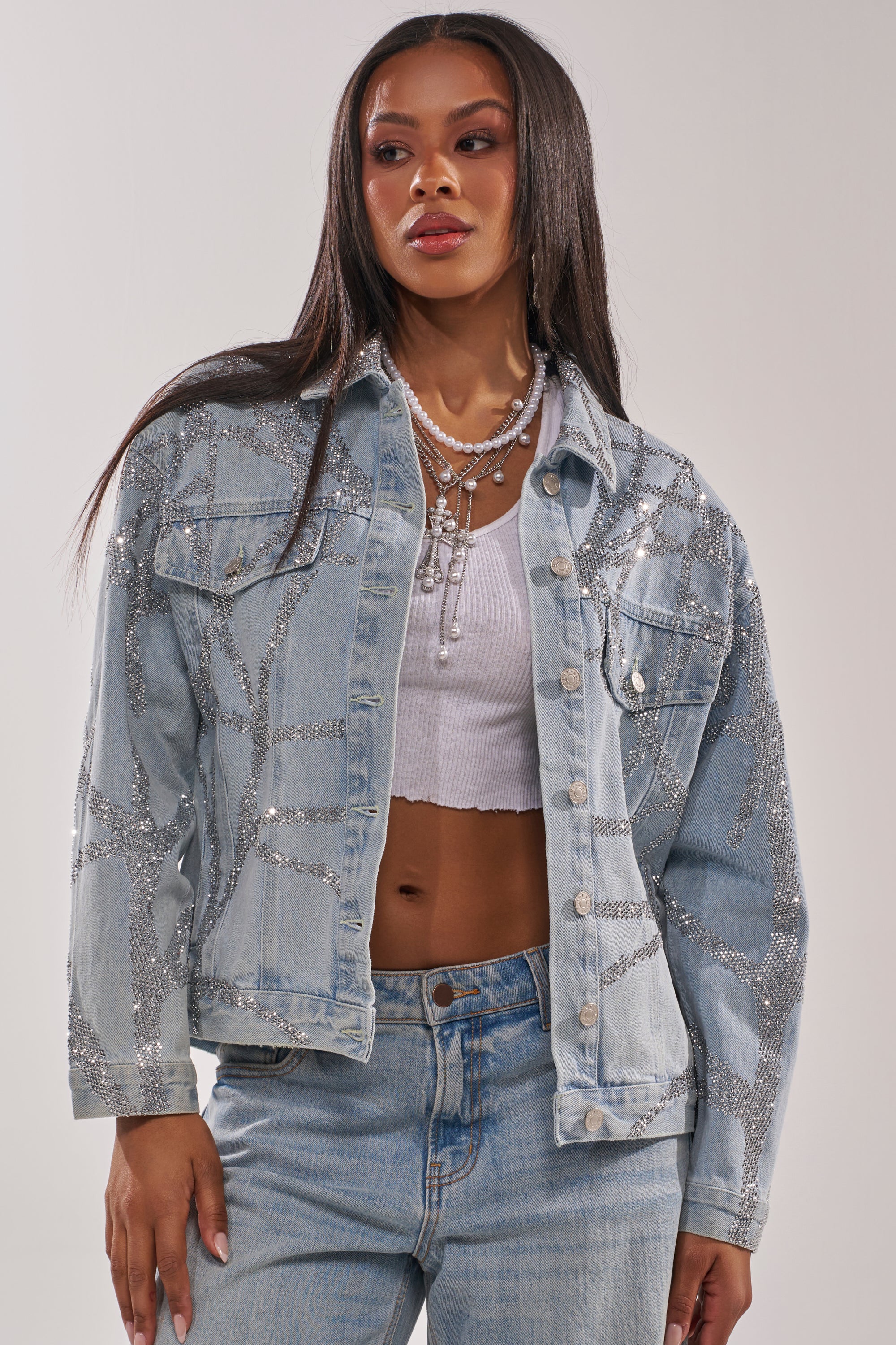 A woman showcases the TWILIGHT ZONE RHINESTONE DENIM JACKET over a white crop top and matching jeans, styled with layered silver necklaces. She has long straight hair and radiates confidence. 2