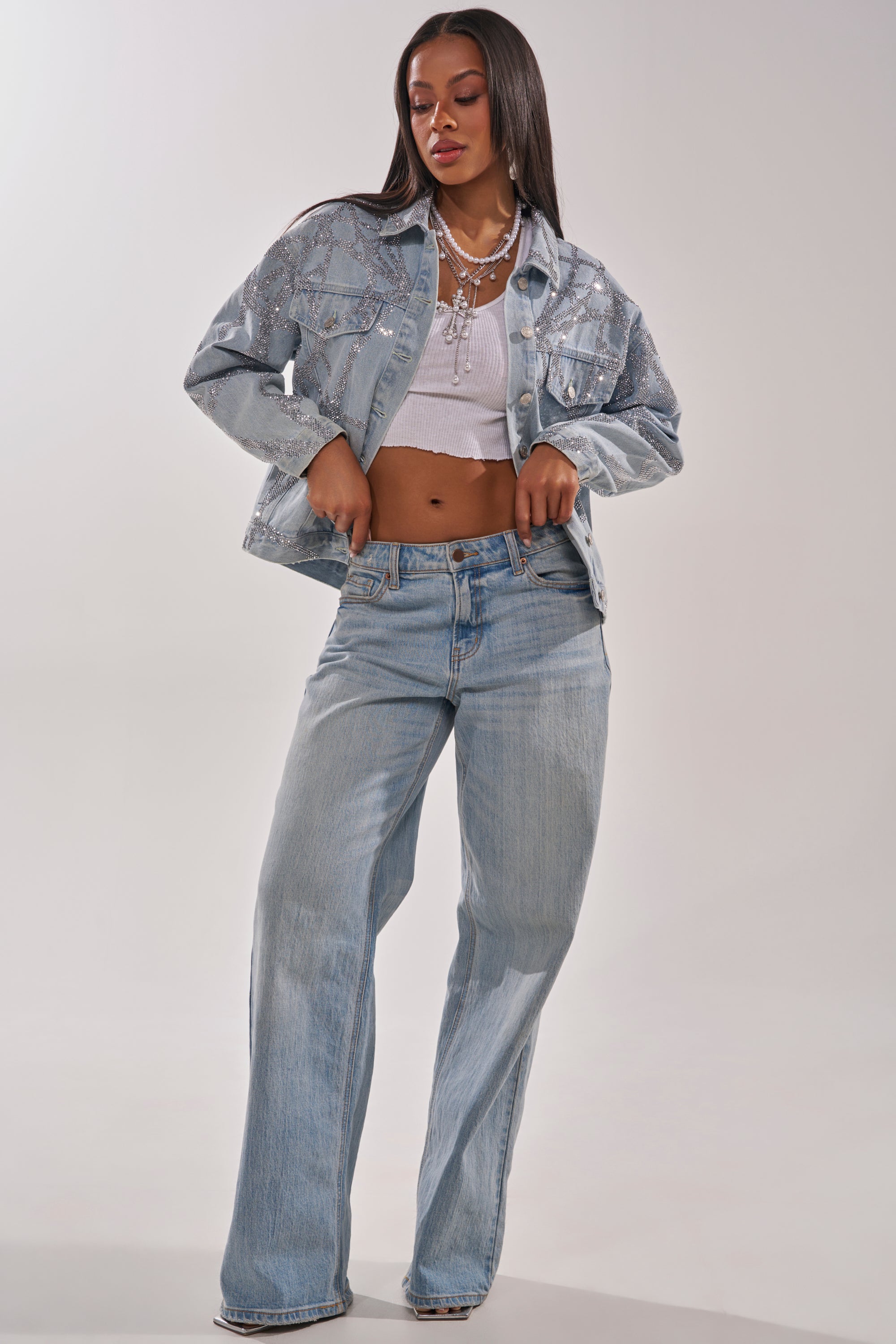 A woman stands confidently wearing the TWILIGHT ZONE RHINESTONE DENIM JACKET, paired with a white crop top, light blue wide-leg jeans, and layered necklaces. She has straight long hair and poses against a neutral background. 7