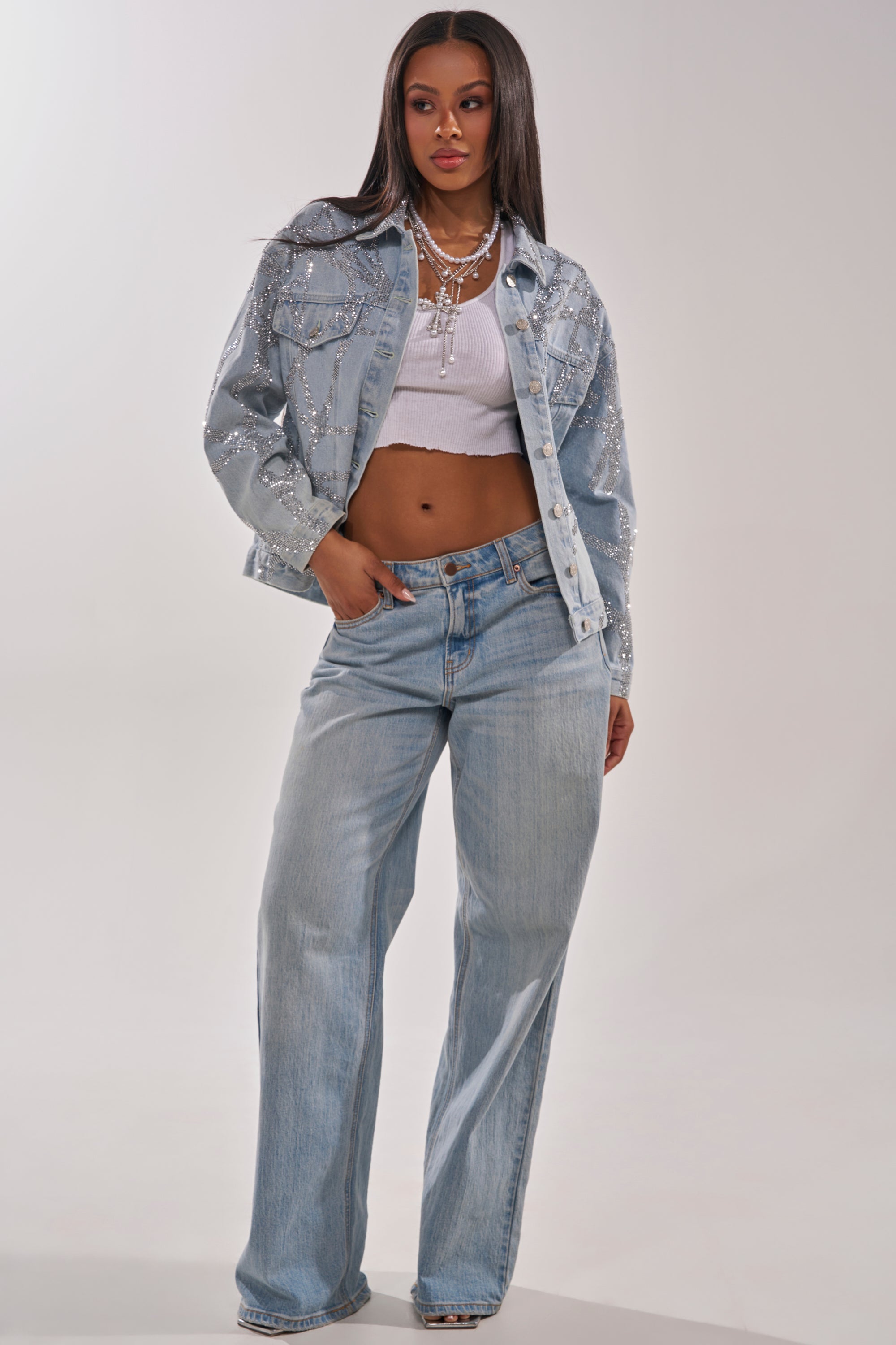 A woman exudes confidence in the TWILIGHT ZONE RHINESTONE DENIM JACKET over a white crop top and loose light blue jeans. Layered necklaces and sleek, long straight hair complete her look against a plain background. 8