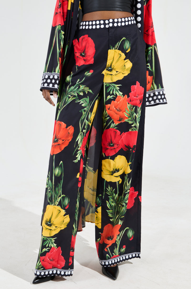 A person wearing BELLADONNA SATIN PALAZZO PANTS featuring a vibrant floral graphic print in shades of red, yellow, and orange. The wide-leg silhouette is enhanced by a black and white polka dot trim, and the ensemble is beautifully complemented by a matching floral cloak.