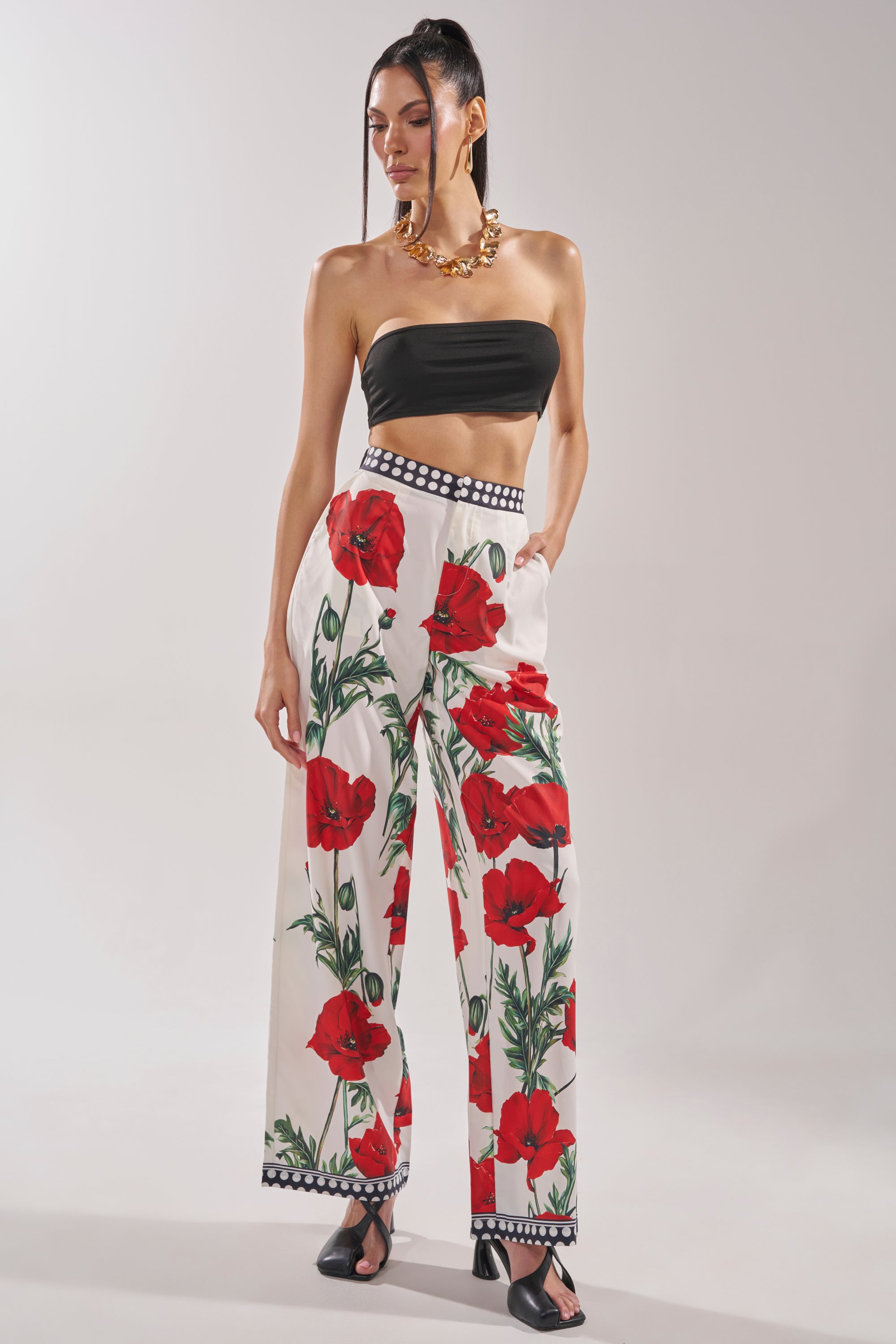 A woman in a black strapless crop top, BELLADONNA PRINT PALAZZO PANTS IN WHITE MULTI, black sandals, and a chunky gold necklace stands against a plain light background.