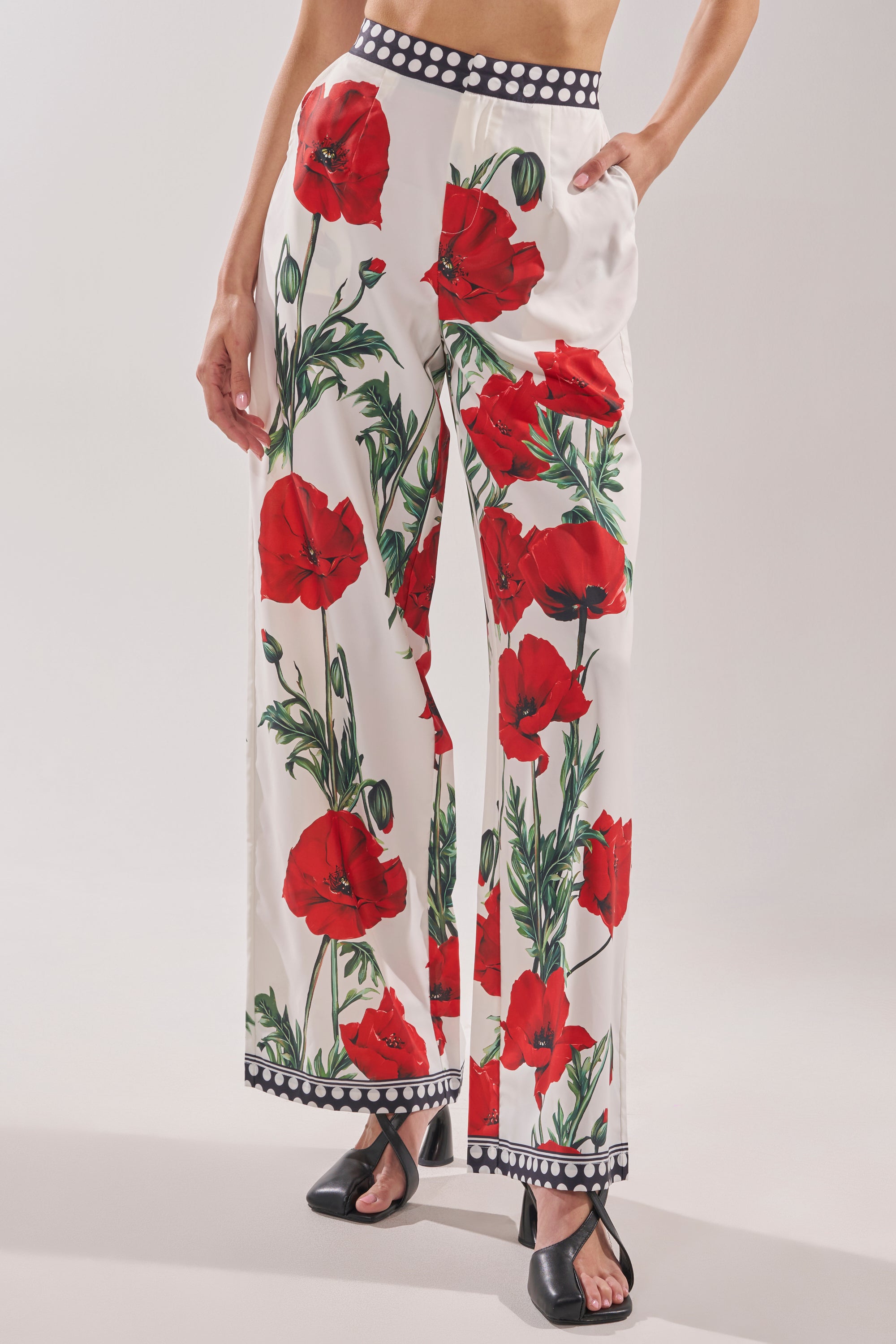 Wearing the BELLADONNA PRINT PALAZZO PANTS IN WHITE MULTI with bold red poppies, green stems, and a polka dot waistband, paired with black open-toe sandals, creates a chic and stylish look.