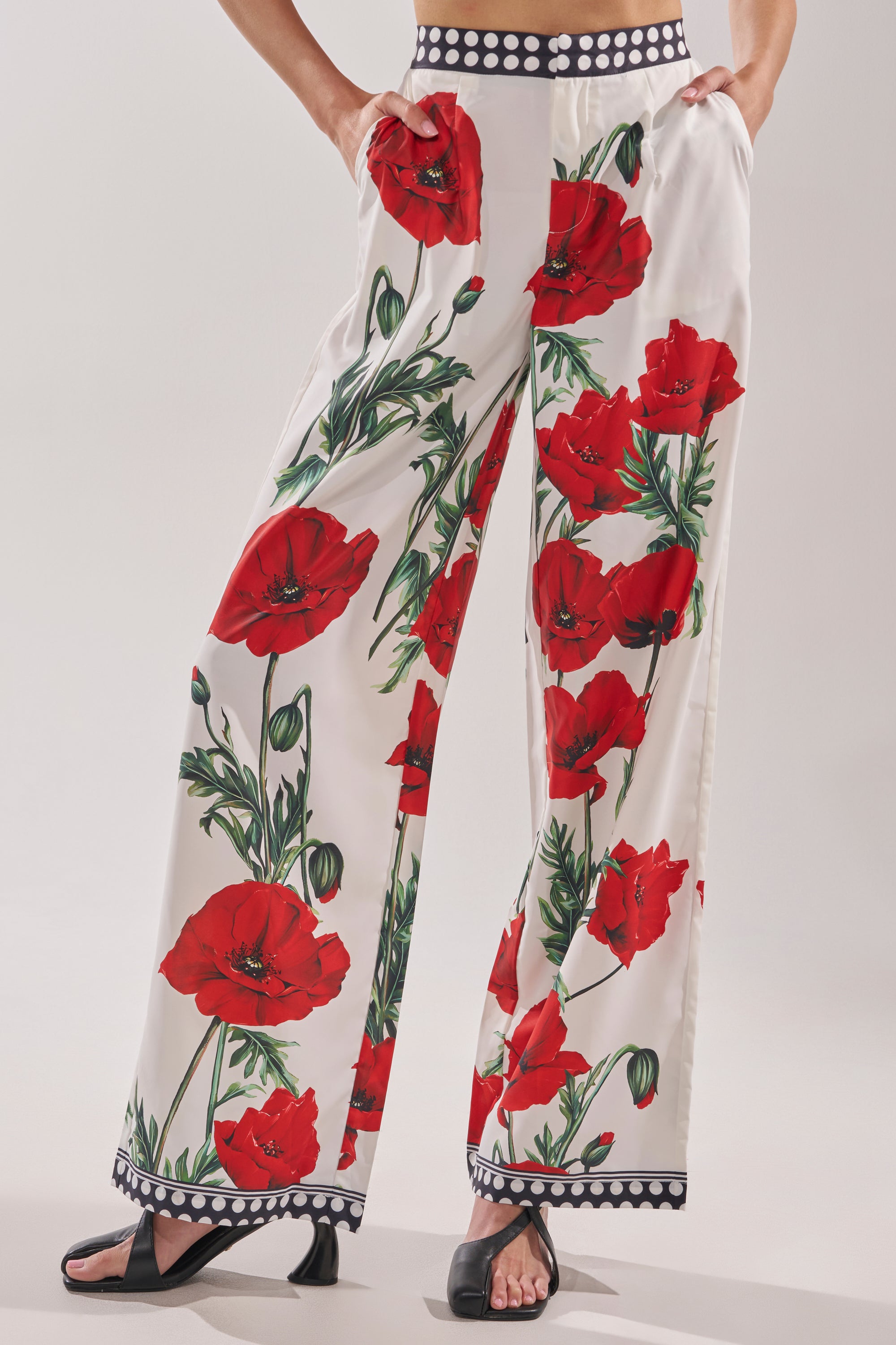 A person models the BELLADONNA PRINT PALAZZO PANTS IN WHITE MULTI, featuring wide legs with bold red poppy florals and black-and-white polka dot waistband and hem, styled with black open-toe sandals against a plain light background.
