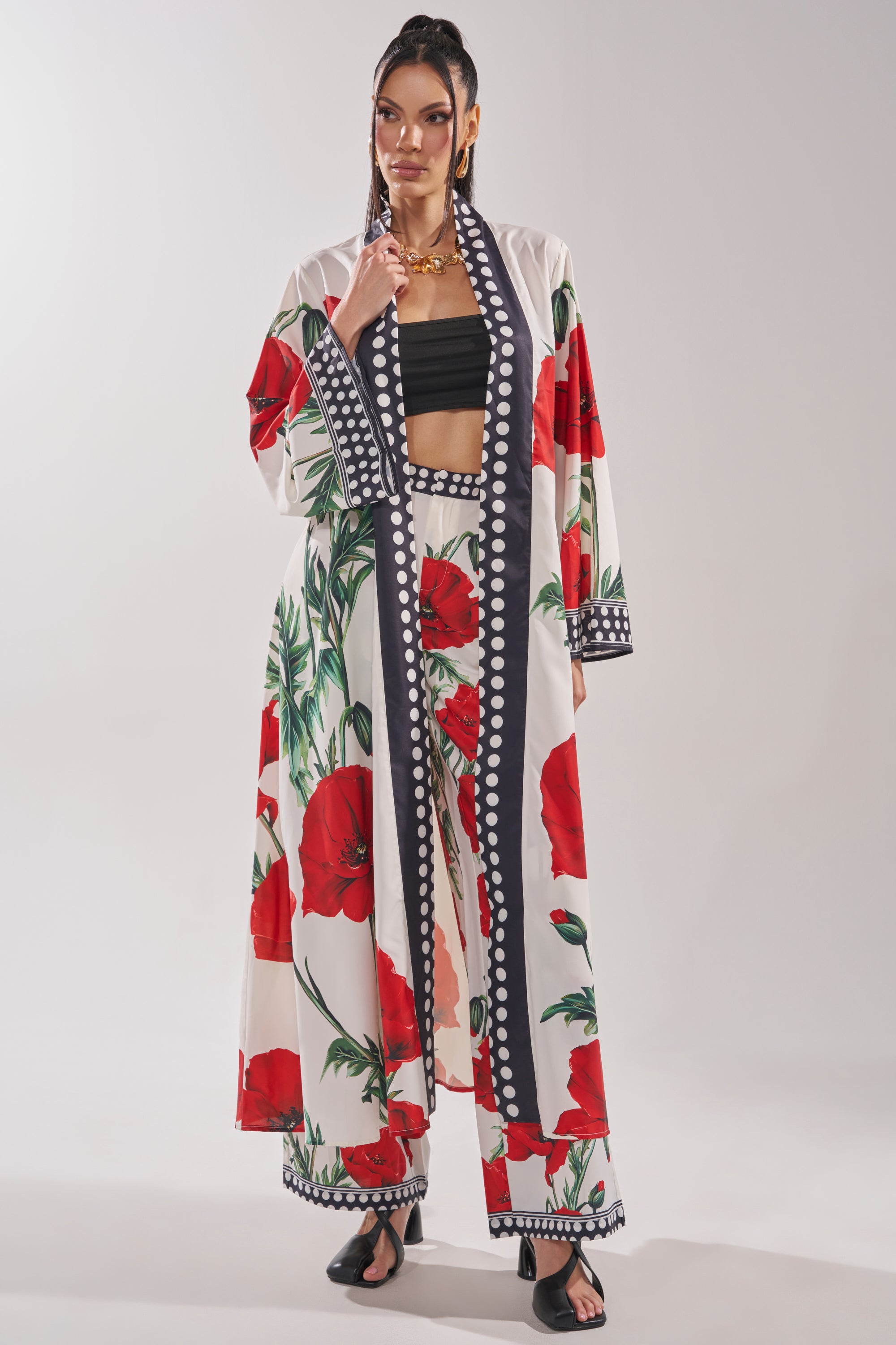 A woman poses against a plain light background wearing the BELLADONNA CHIFFON DUSTER and matching wide-leg pants with bold red poppy prints, paired with a black crop top, black sandals, and gold jewelry.