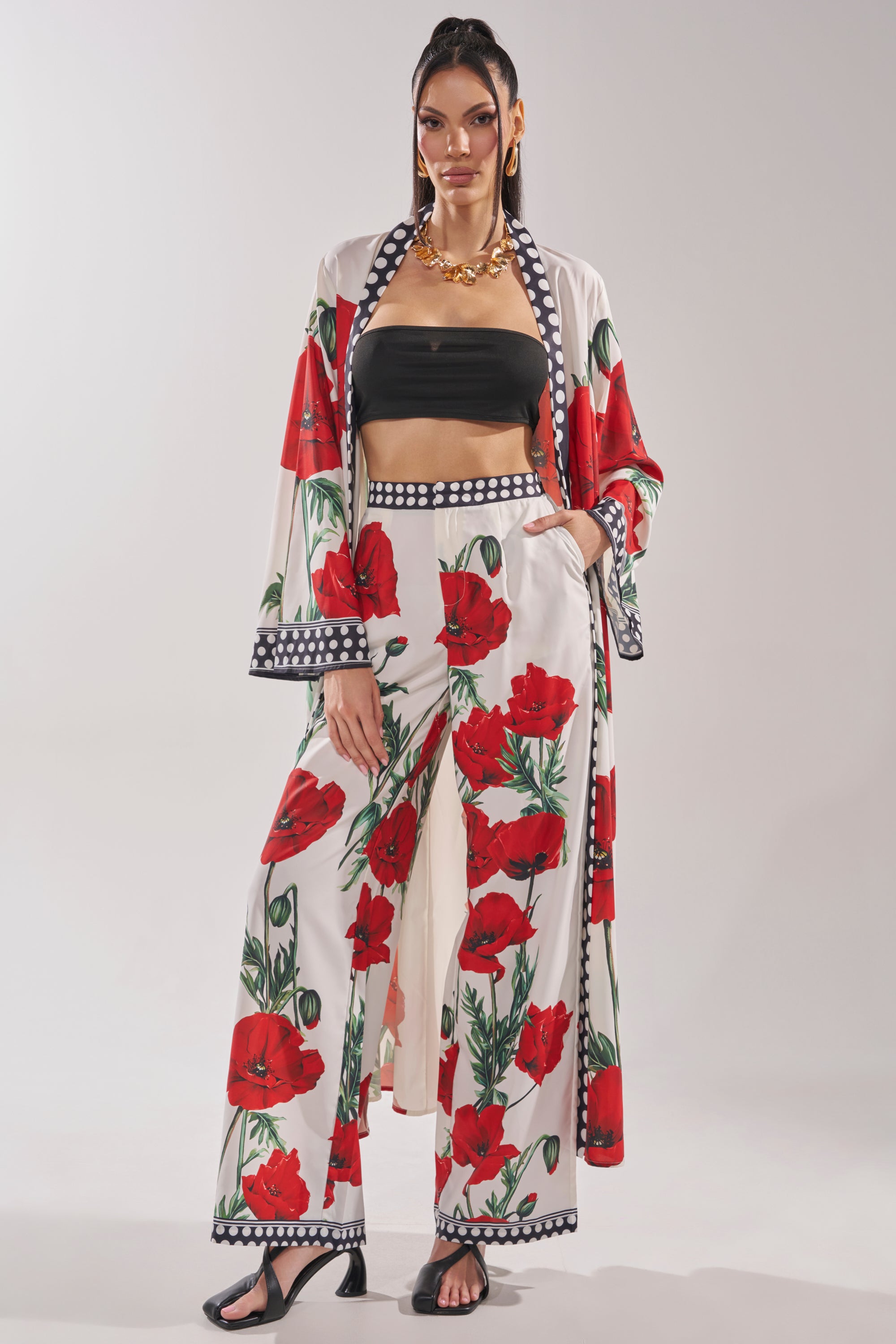 A woman poses in a coordinated outfit featuring wide-leg floral pants, a black bandeau top, gold chunky jewelry, black sandals, and the BELLADONNA CHIFFON DUSTER against a plain background.