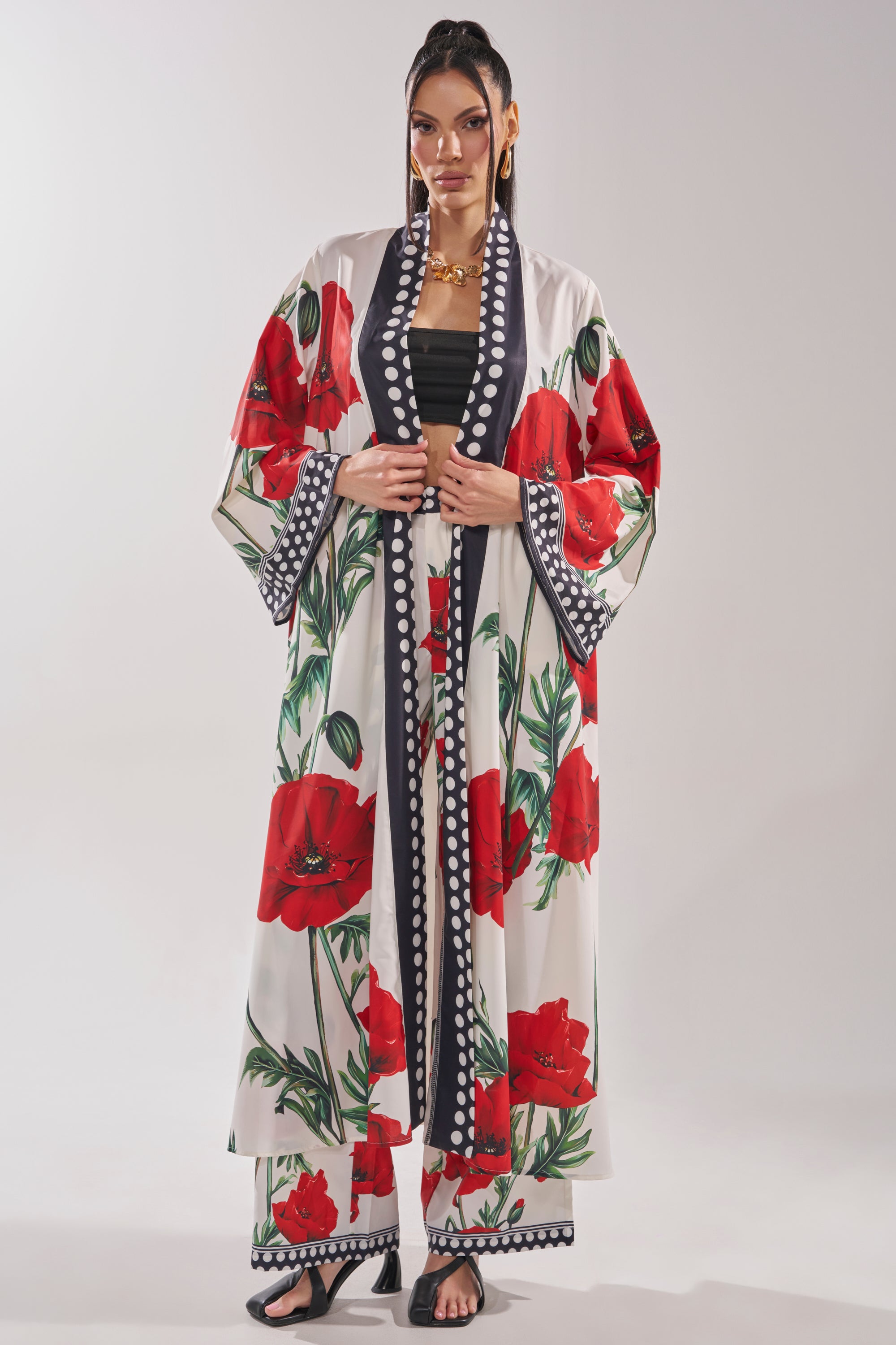 A woman wears the BELLADONNA CHIFFON DUSTER, featuring a vintage-inspired floral print with black and white polka dot trim, paired with matching pants, black sandals, and gold jewelry for a striking longline silhouette against a plain light background.
