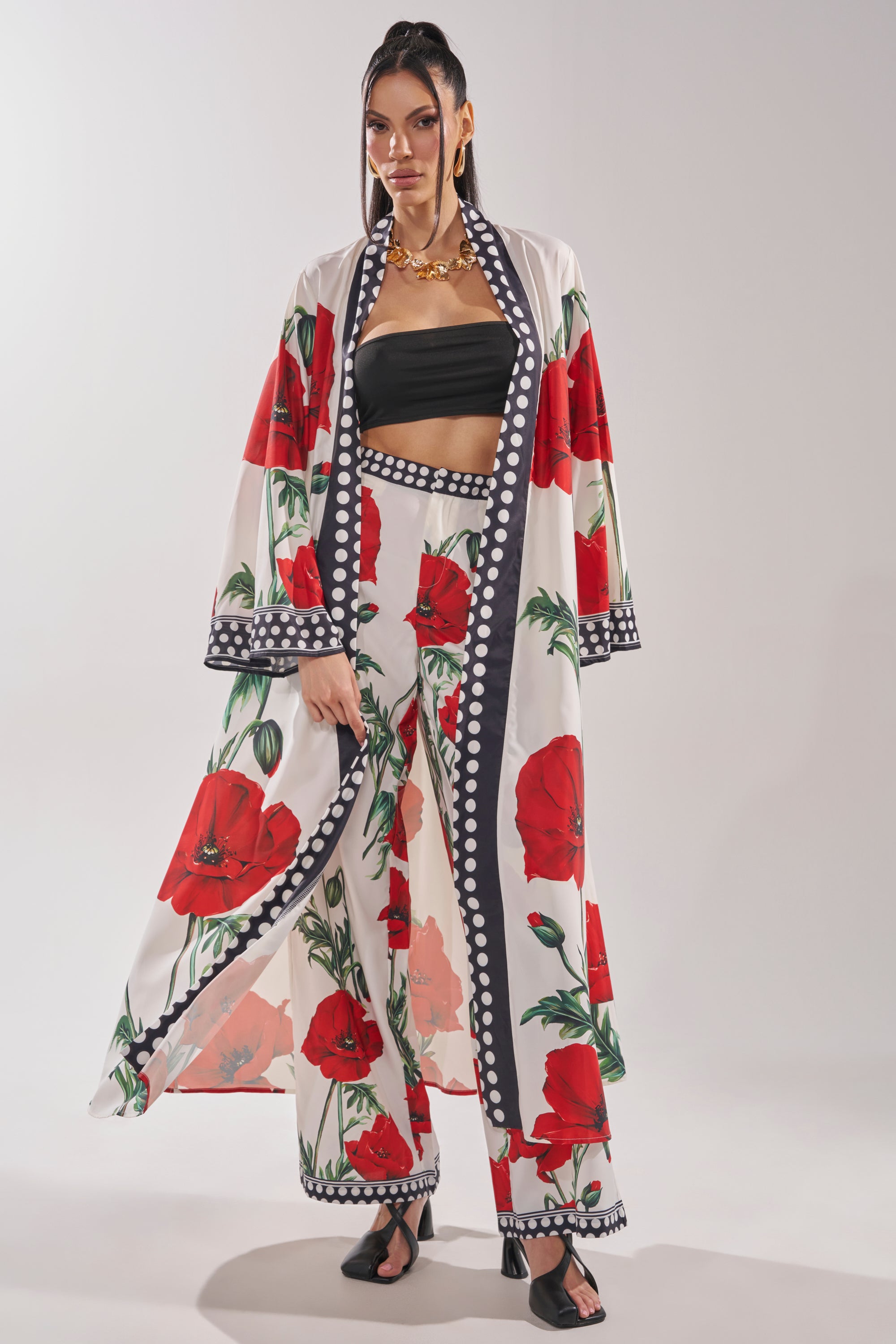 A woman wears the BELLADONNA CHIFFON DUSTER—a white longline kimono with large red floral prints—paired with matching wide-leg pants, a black bandeau top, black sandals, and a gold necklace against a plain backdrop.
