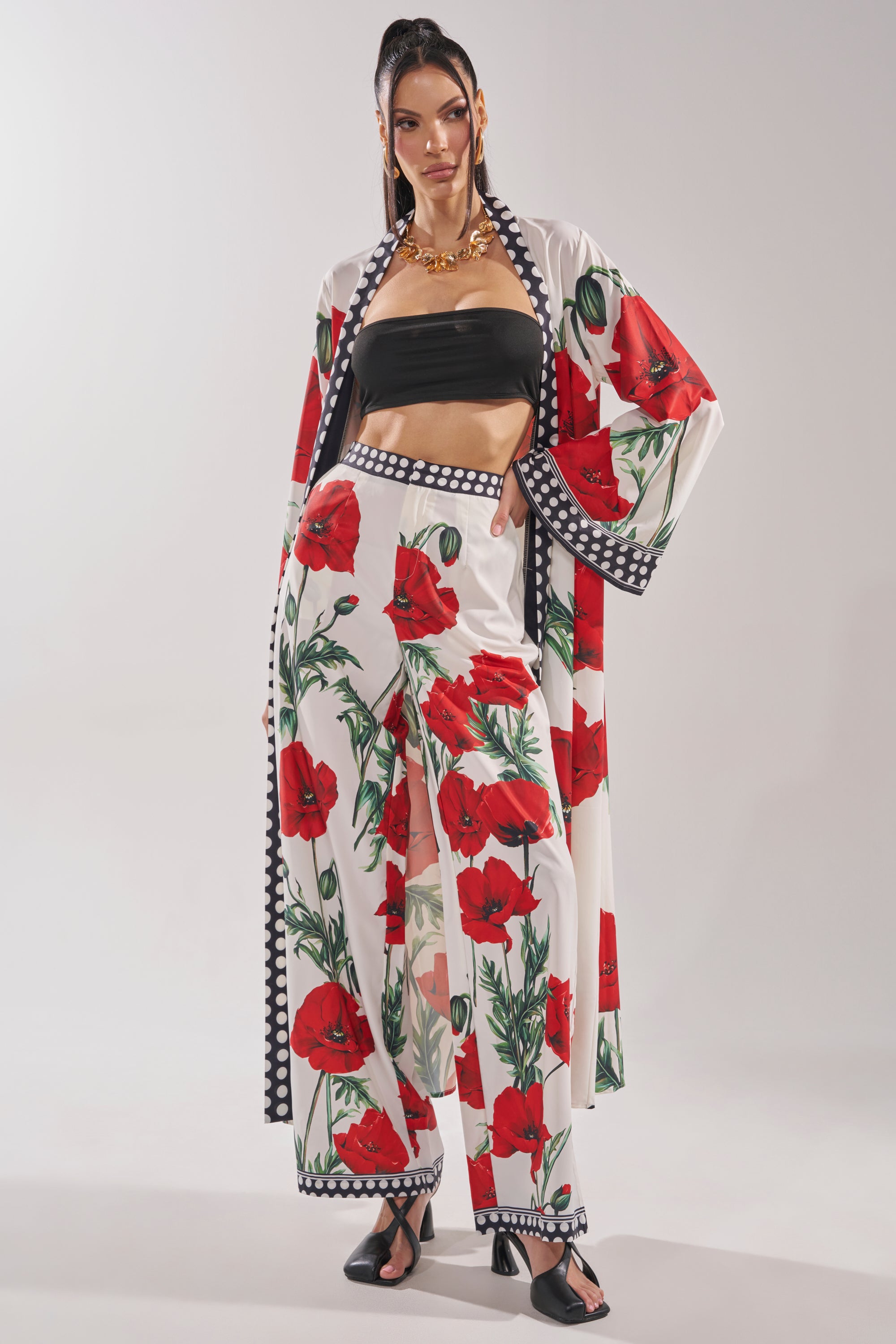 A woman models the BELLADONNA CHIFFON DUSTER—a white pantsuit with a bold red floral print and black-and-white trim—paired with a black bandeau top, black sandals, gold jewelry, and a high ponytail.