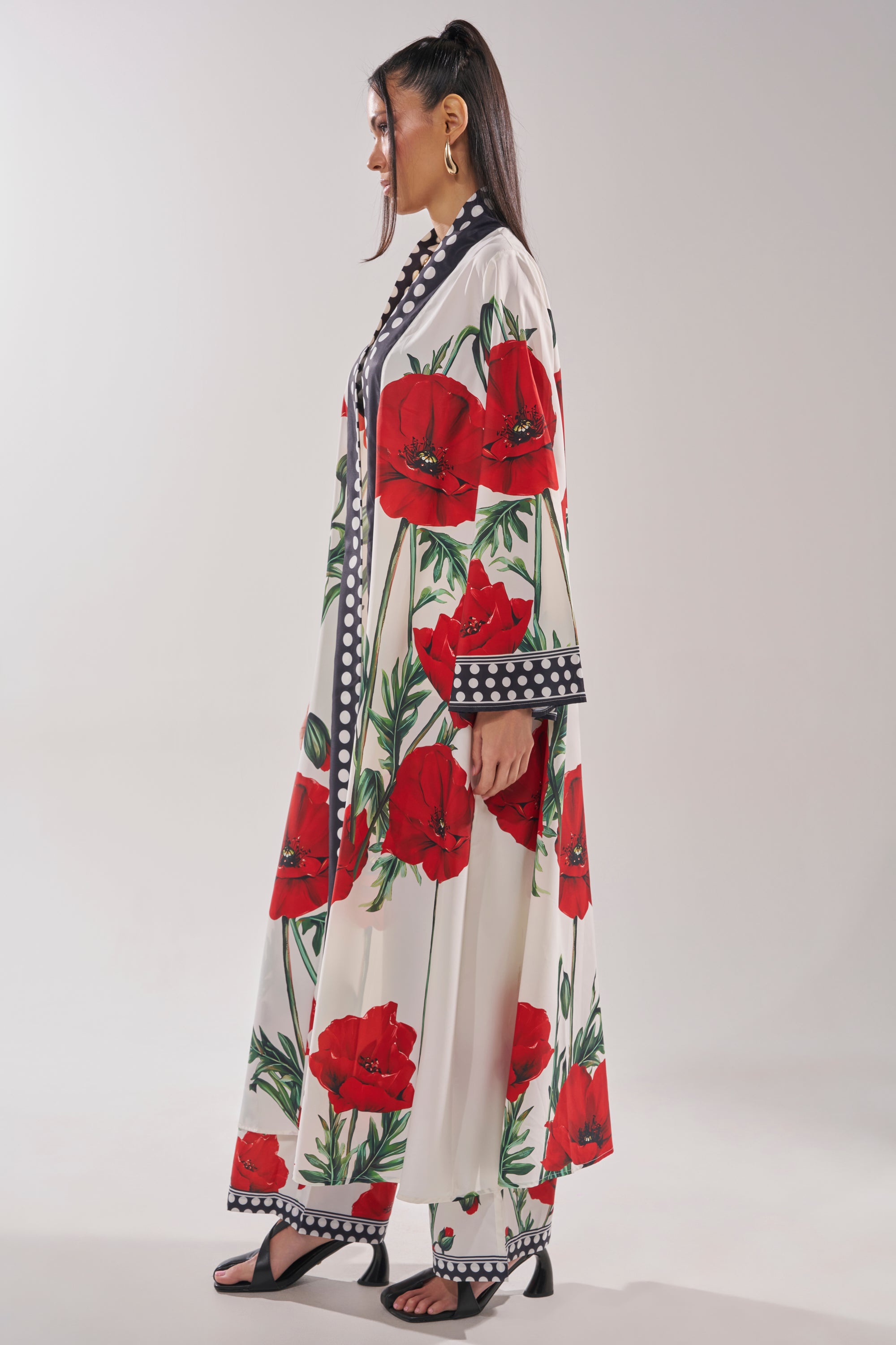 A woman in profile wears the BELLADONNA CHIFFON DUSTER with bold red poppy prints, matching wide-leg pants, hoop earrings, and black sandals against a plain background.