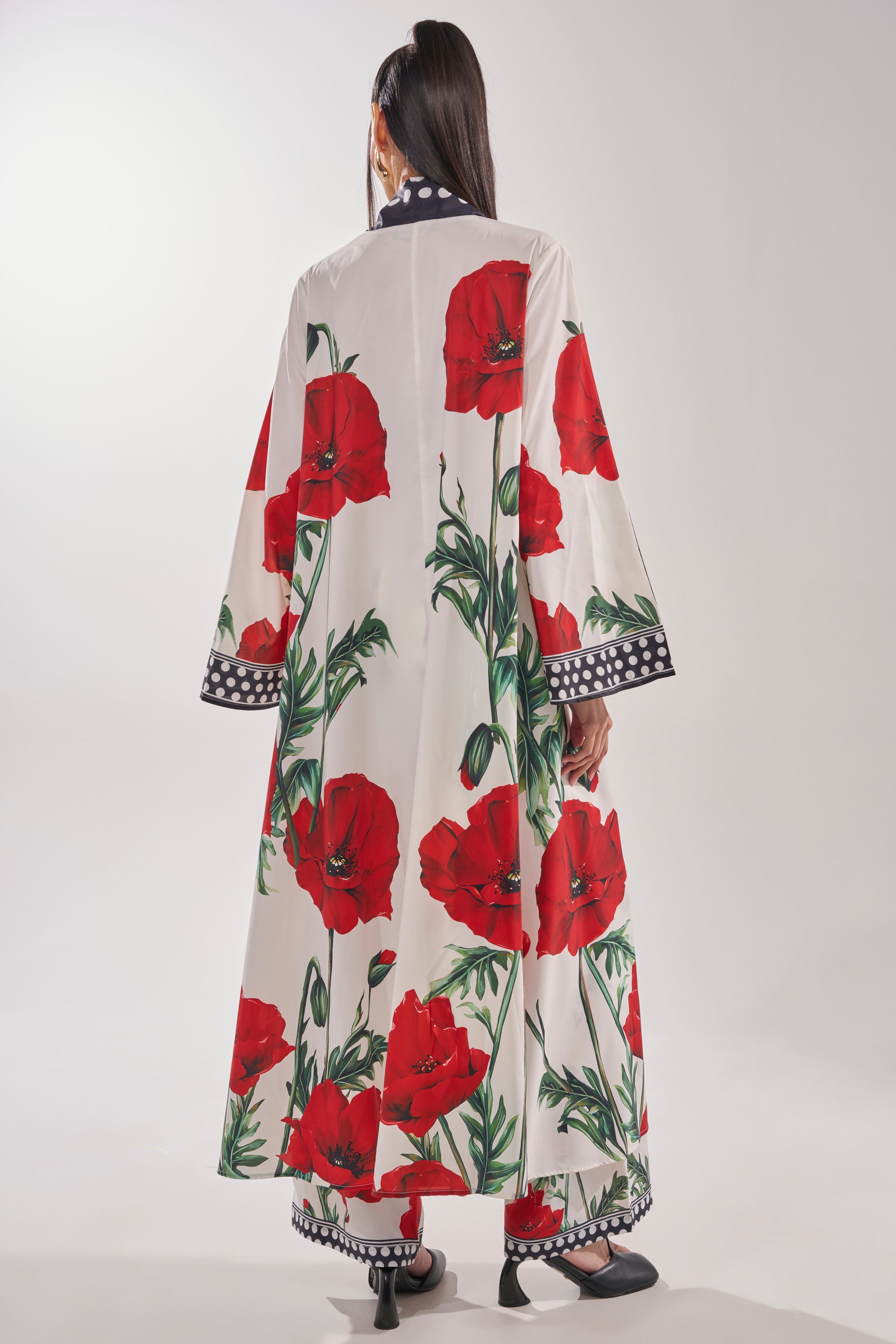 A woman faces away wearing the BELLADONNA CHIFFON DUSTER, a long white dress with bold red poppy prints and black-and-white polka dot trim, paired with matching pants and black high-heeled sandals.